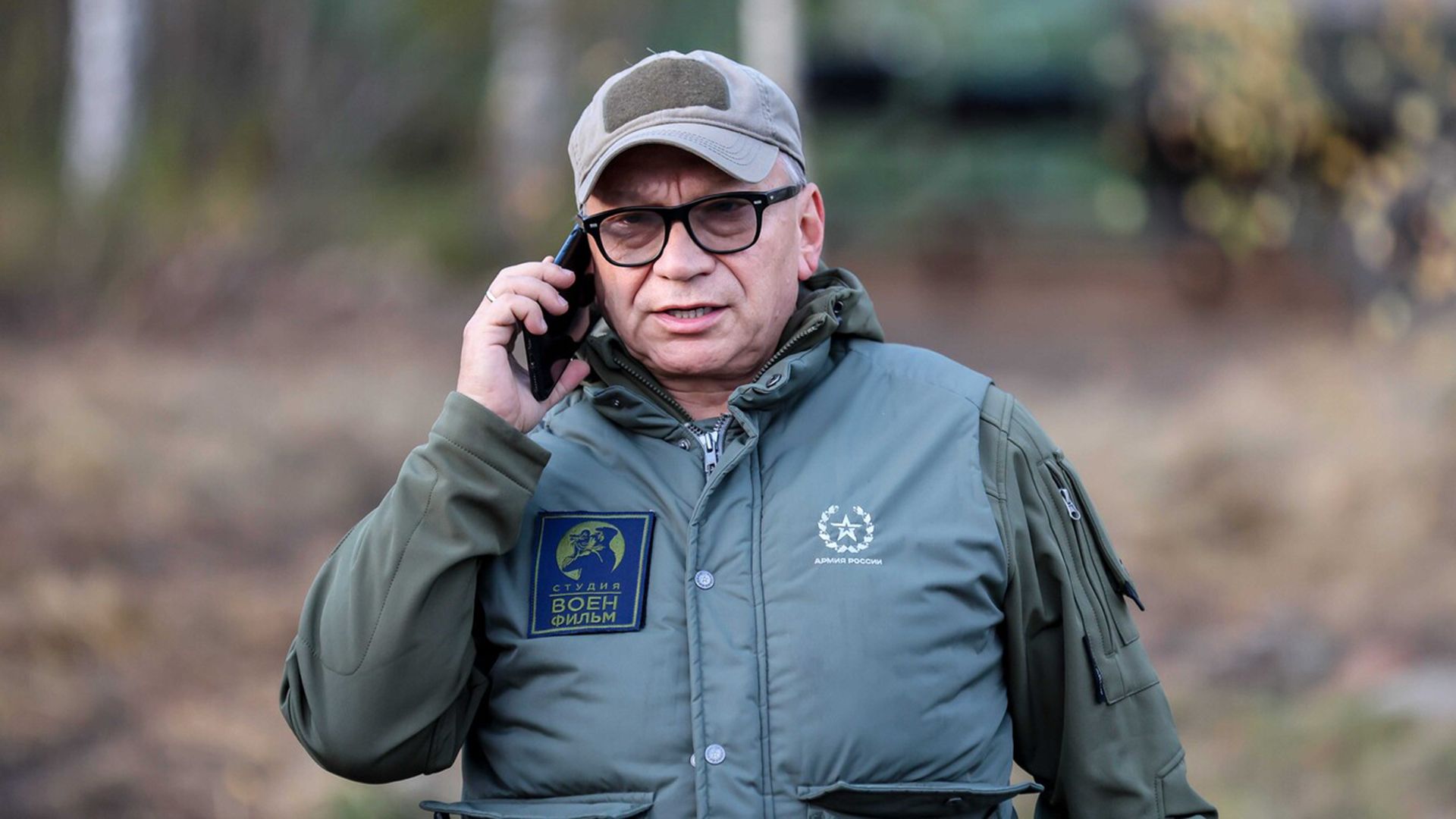 Игорь Угольников в куртке цвета хаки и кепке разговаривает по смартфону
