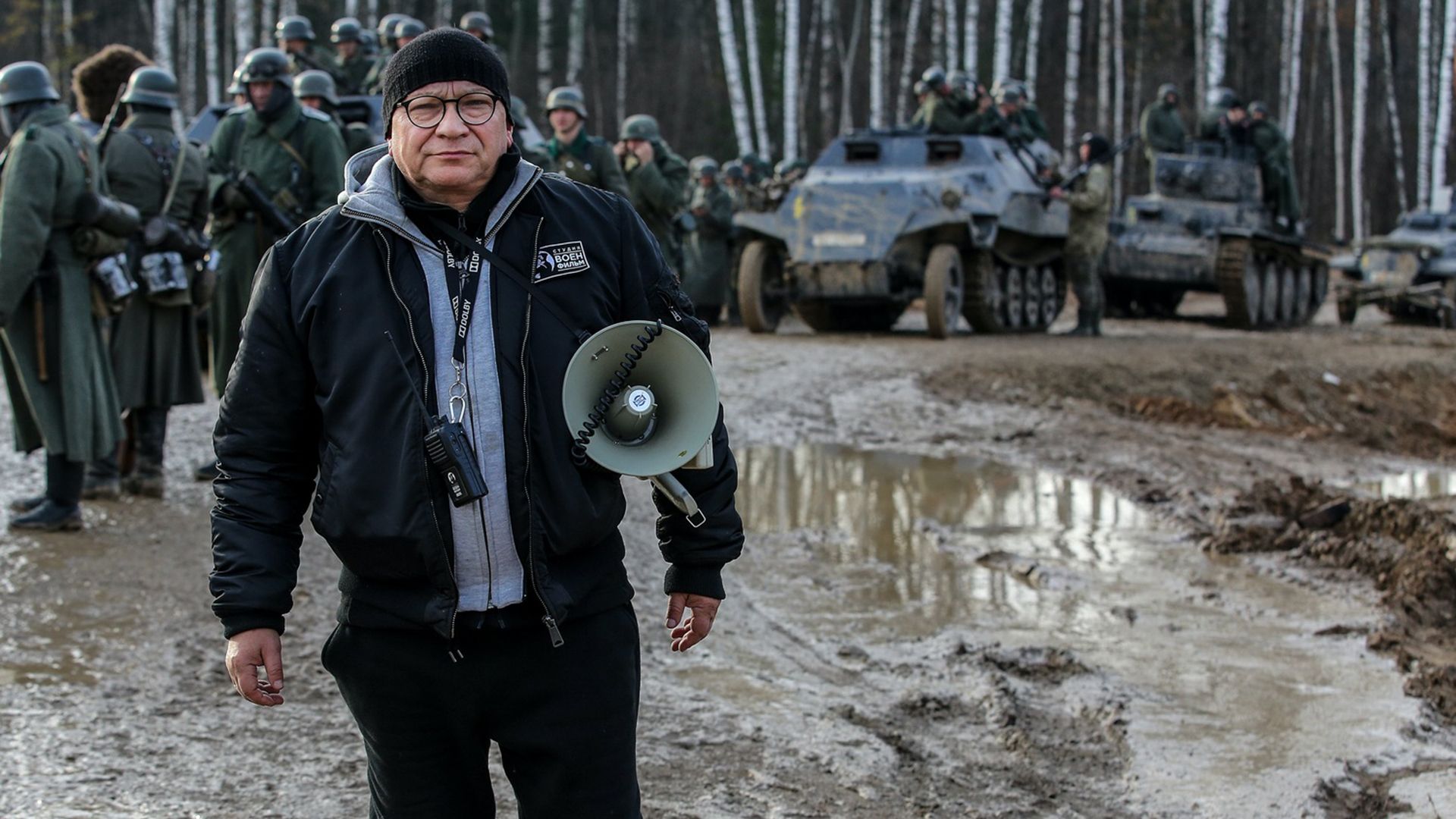 Игорь Угольников с мегафоном подмышкой стоит на грязной тропе на фоне танков и артистов в немецкой военной форме времен ВОВ