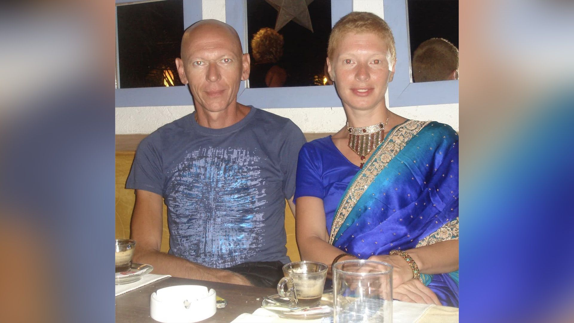 Амалия Мордвинова с остриженными волосами и в сари сидит за столом с лысым мужчиной в серой футболке