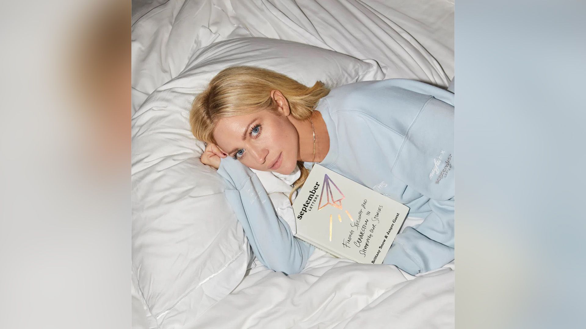 Бриттани Сноу в голубой толстовке лежит на белых простынях с книгой "Сентябрьские письма"