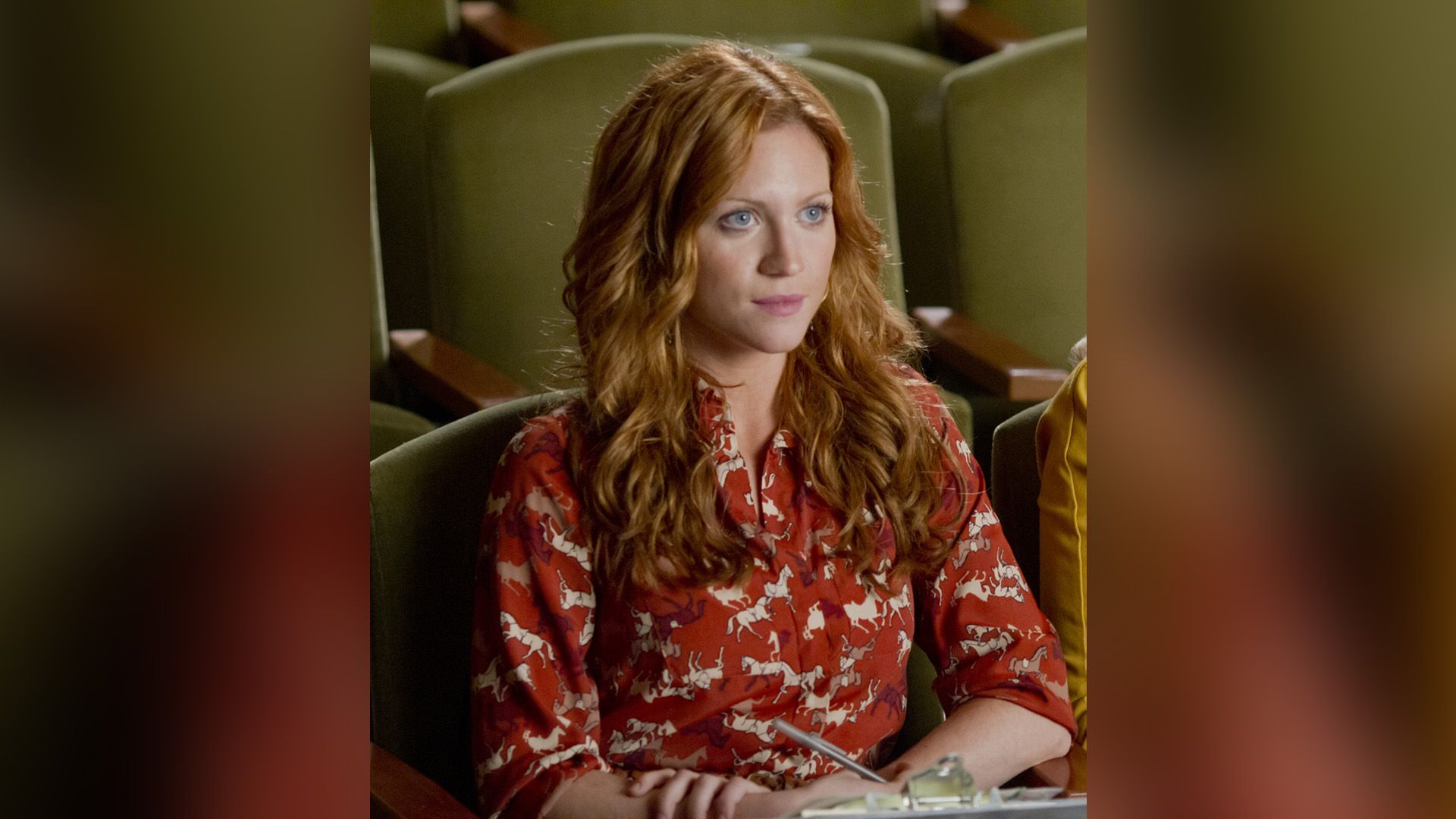 Рыжеволосая Бриттани Сноу в красной рубашке с принтом в виде лошадей сидит в кресле зрительного зала