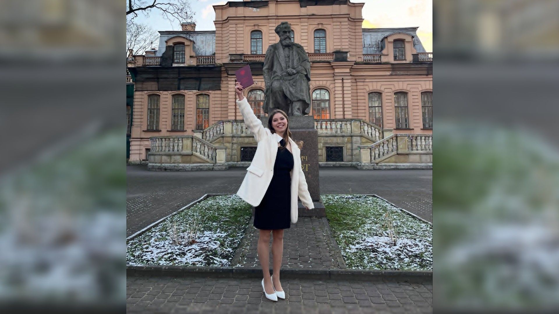 Анастасия Мишина улыбается и держит в руках диплом на фоне здания университета Лесгафта