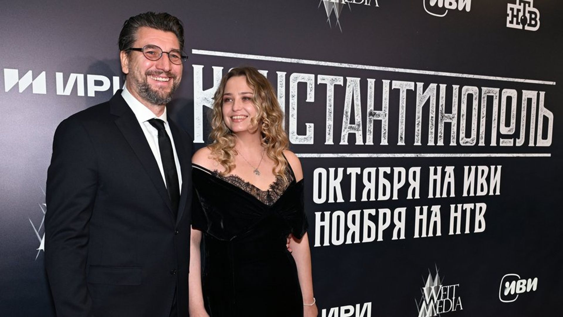 Александр Устюгов и Аглая Шиловская в чёрных нарядах держатся за руки и улыбаются