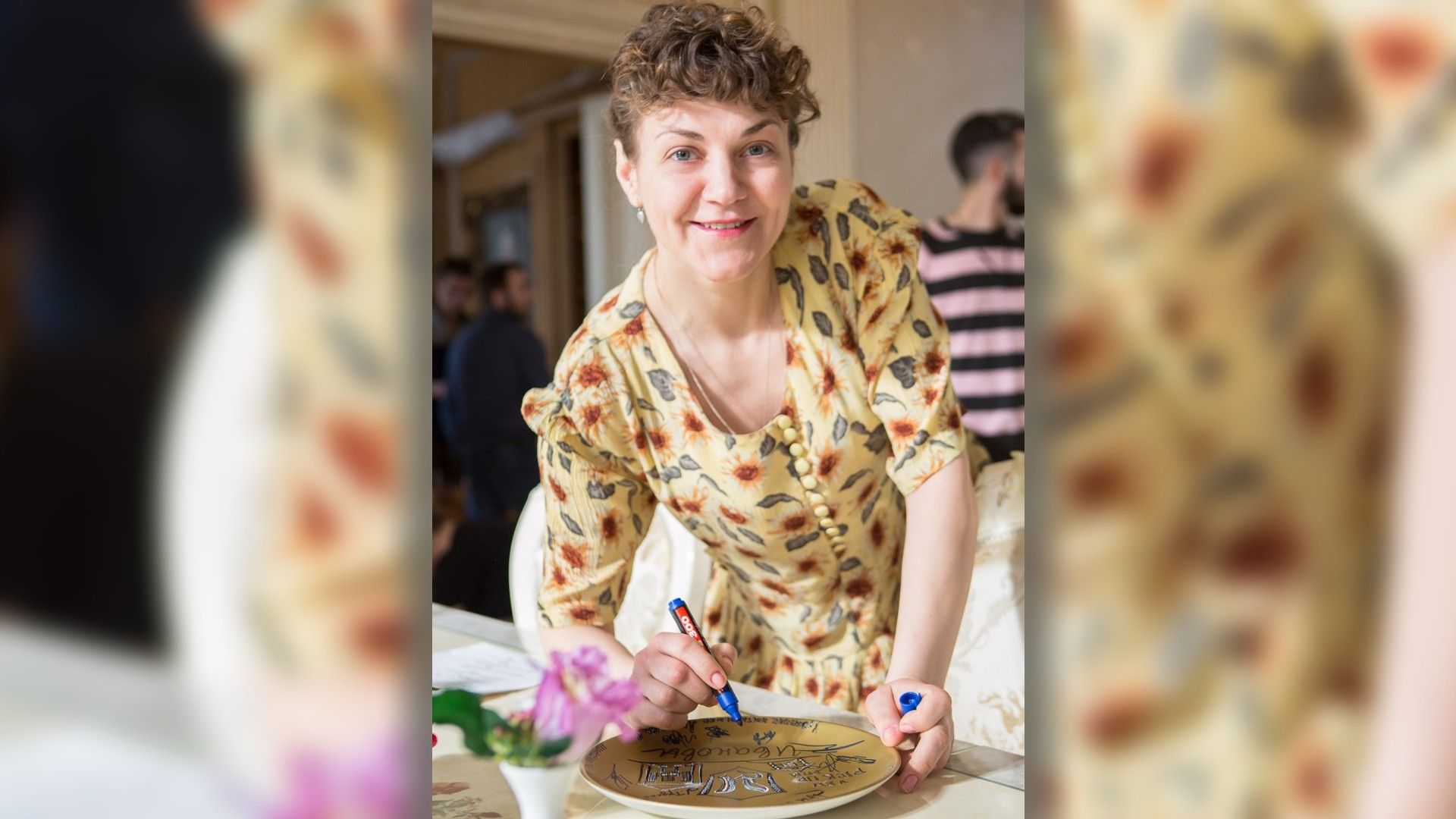 Анна Уколова улыбается, подписывая лежащую на столе тарелку