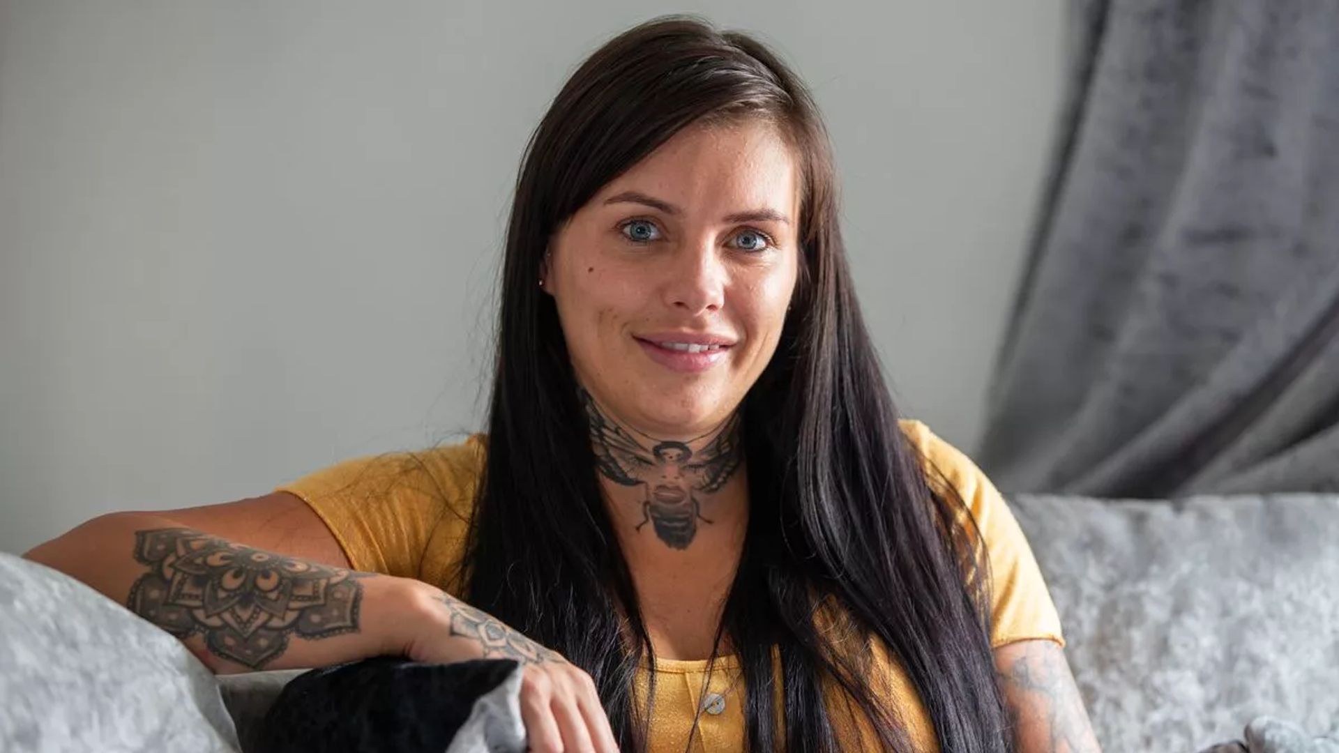 Девушка с длинными черными волосами и татуировками улыбается в камеру