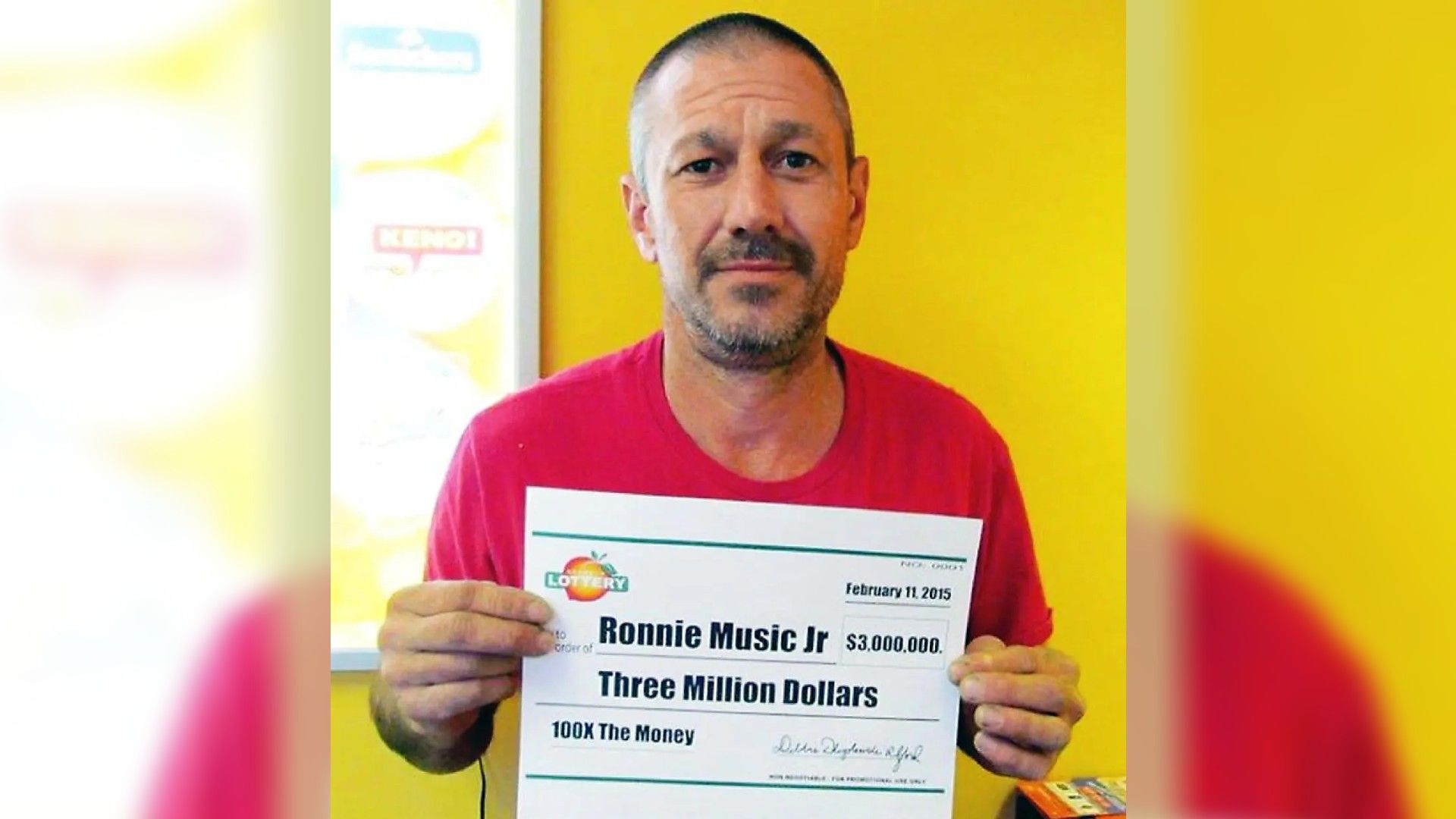 Ронни Мьюзик в красной футболке держит билет с выигрышем 3 миллиона долларов