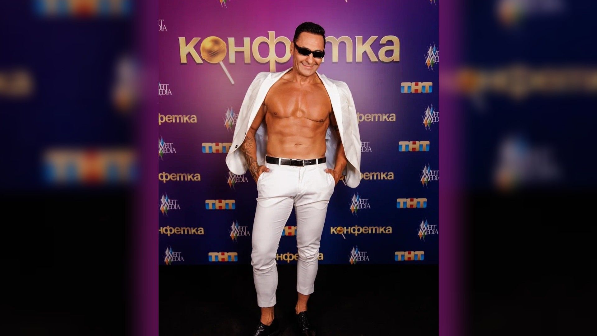 Стас Костюшкин в расстегнутой белой рубашке позирует на фоне надписи Конфетка