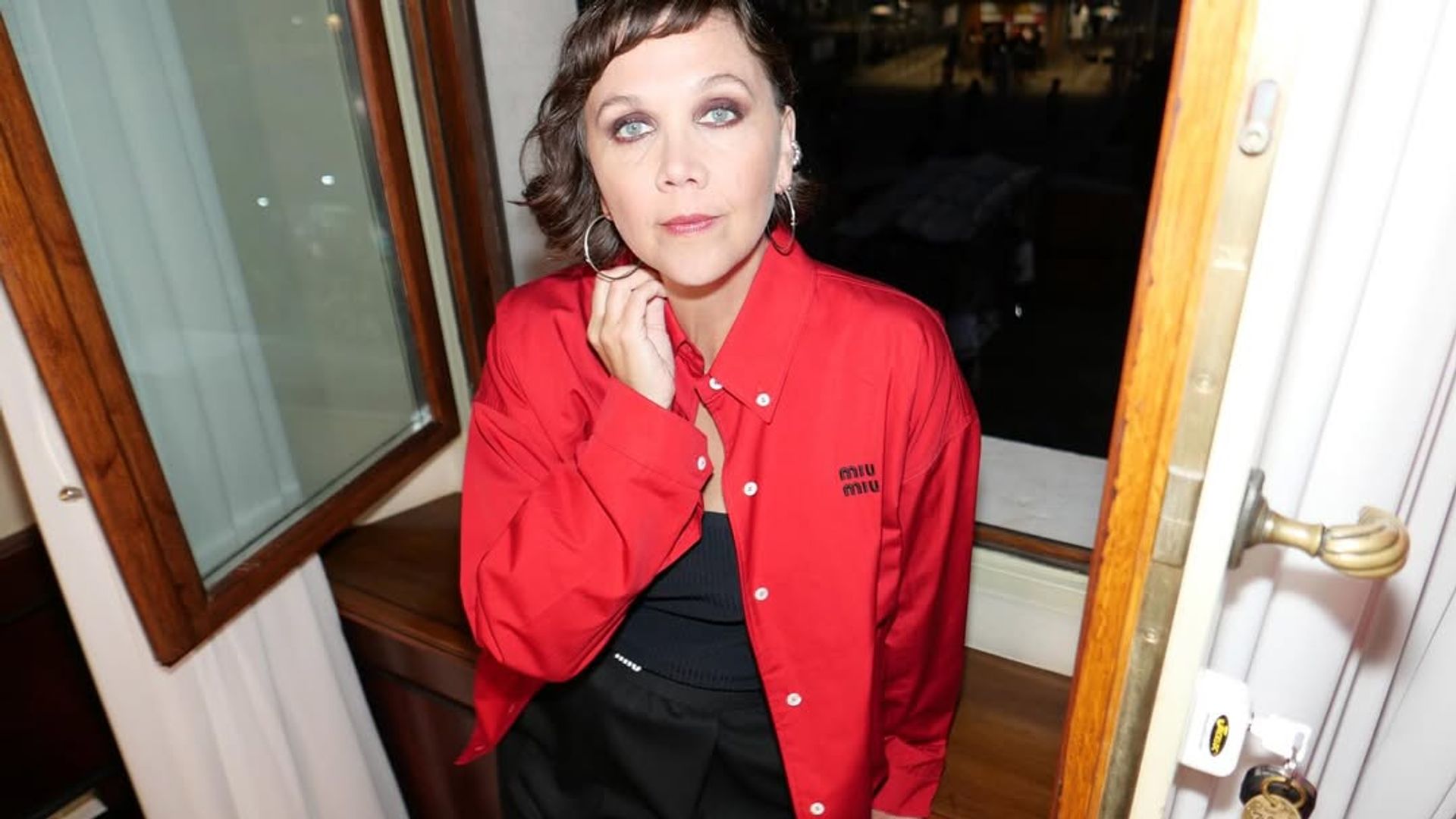 Мэгги Джилленхолл в красной куртке стоит в центре кадра, ракурс сверху