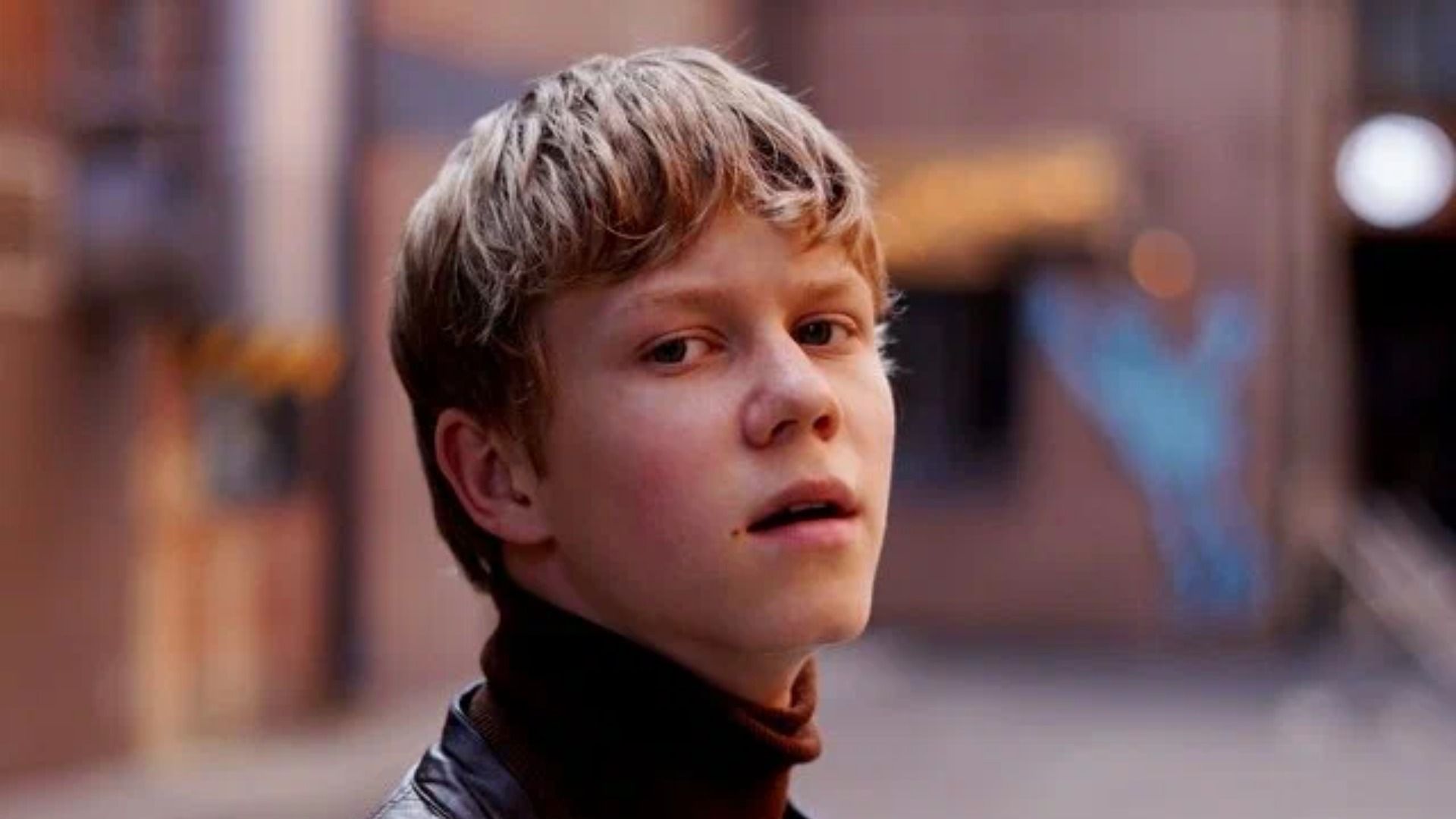 Актер Тимофей Кочнев в черной куртке стоит на фоне размытой улицы