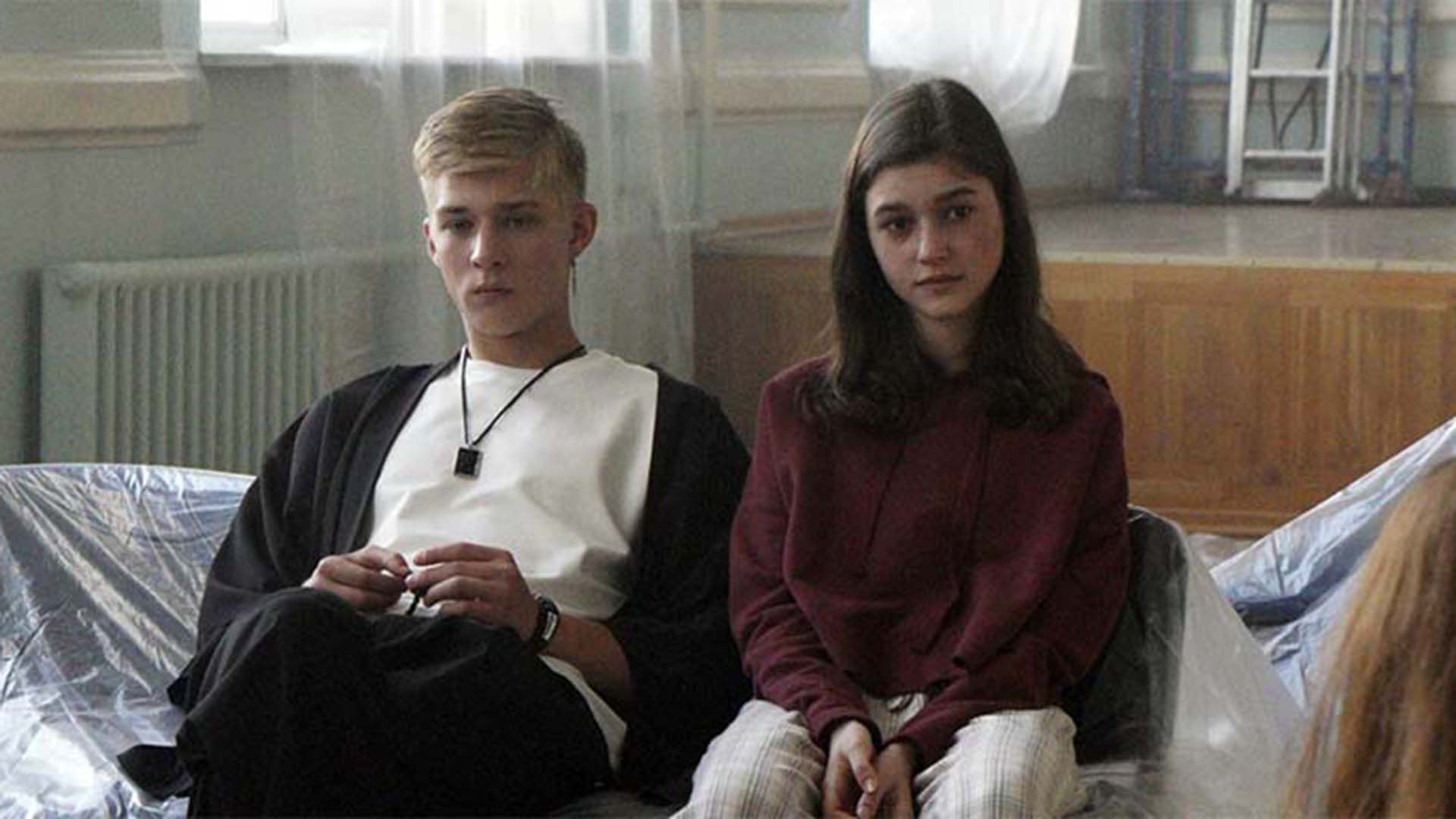 Олег Савостюк и девушка в красном зуди сидят на диване, укрытом плёнкой