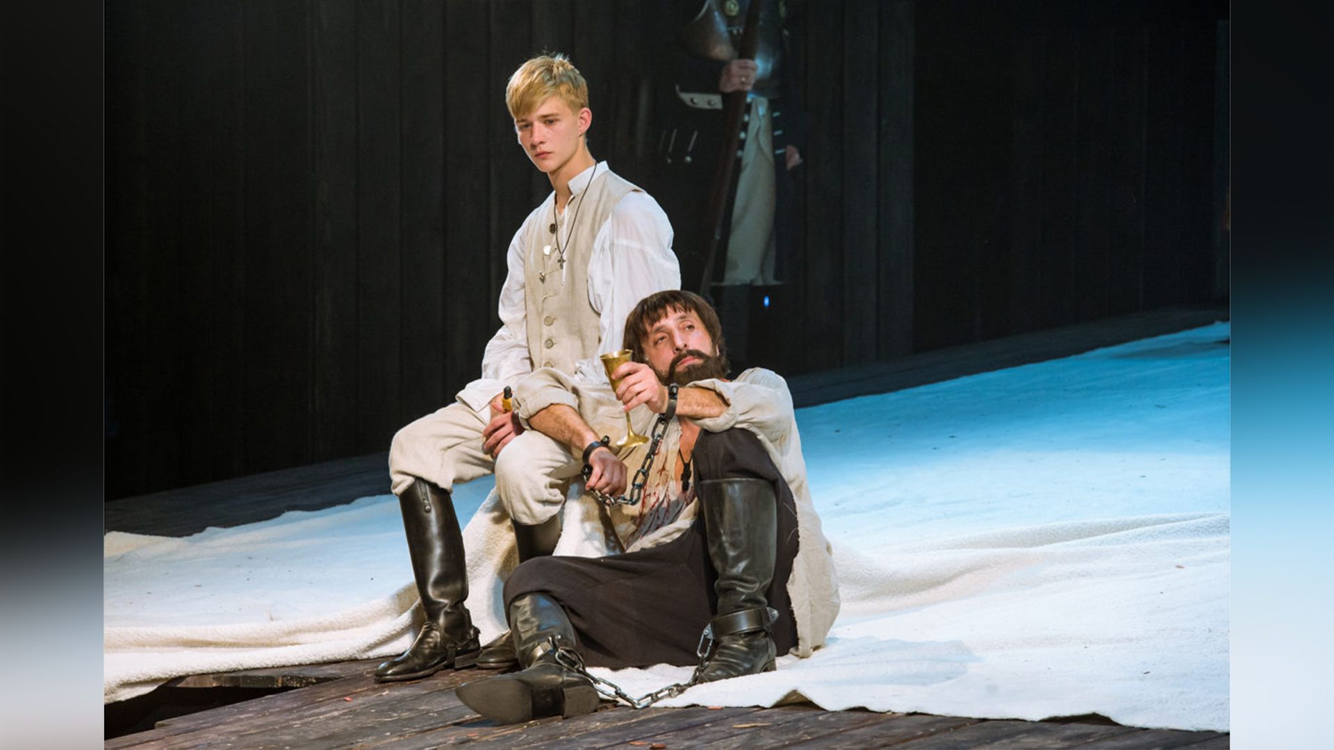 Олег Савостюк в льняных штанах и белой рубашке сидит на стуле на театральной сцене, рядом на полу сидит бородатый мужчина в кандалах