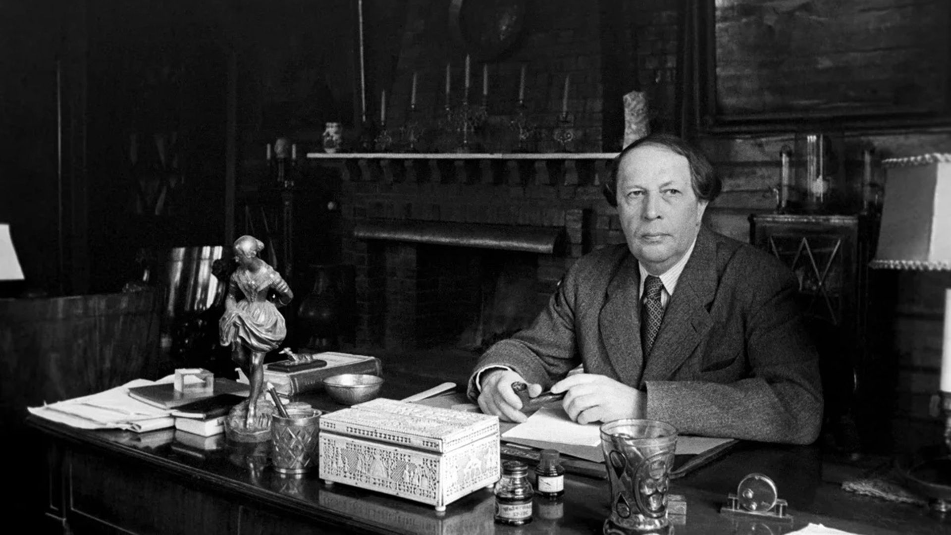 Алексей Толстой сидит за письменным столом, держа в руках трубку. Сзади него большой камин