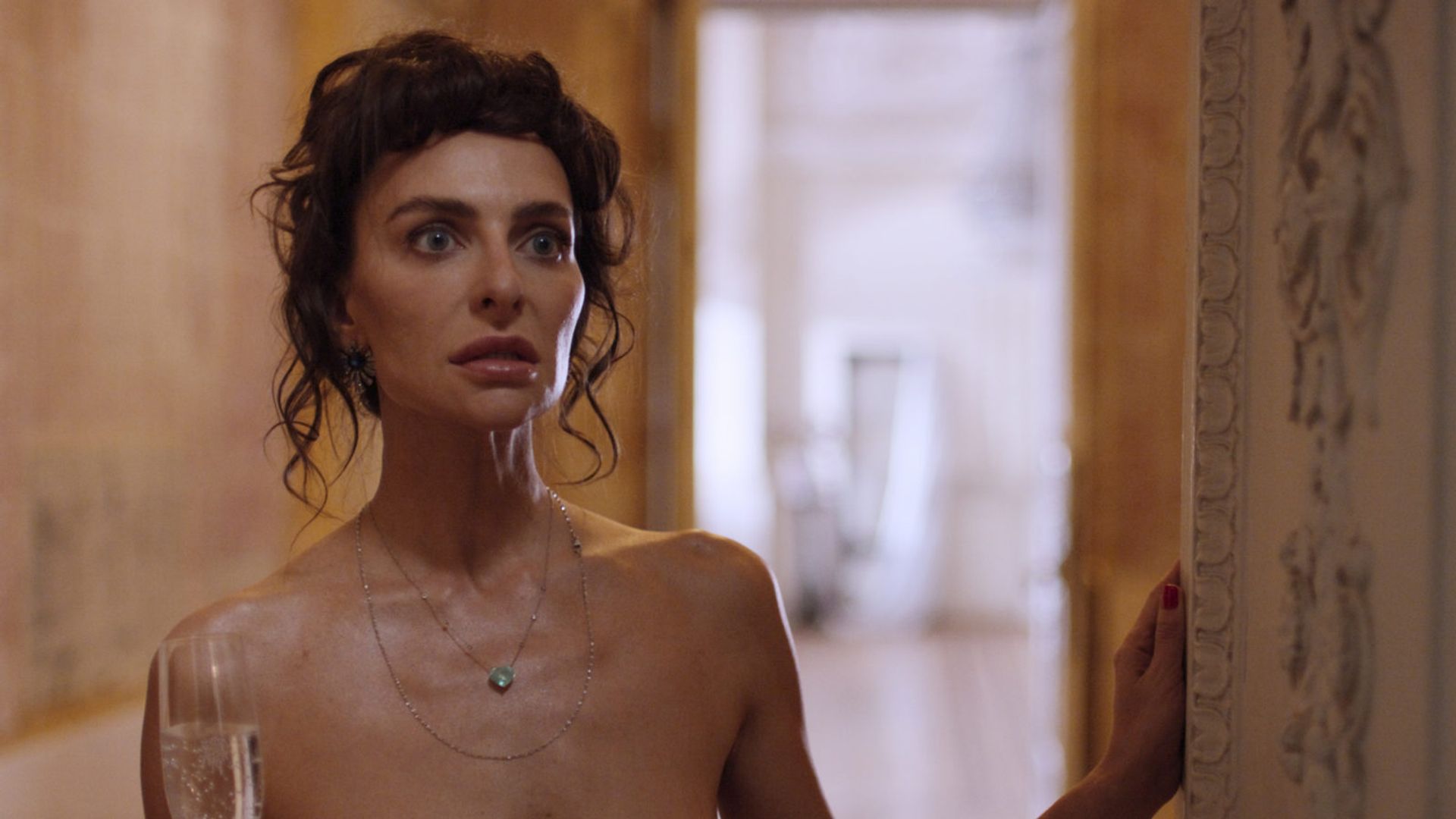 Екатерина Варнава с голыми плечами стоит, опираясь на дверной косяк, с бокалом шампанского в руке