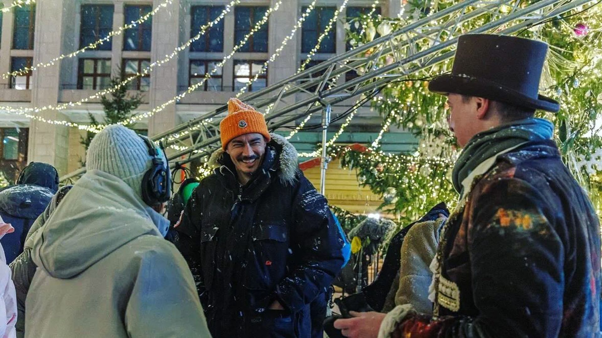 Украшенная новогодними украшениями площадь, идут съемки фильма, в центре мужчина с бородой в оранжевой шапке