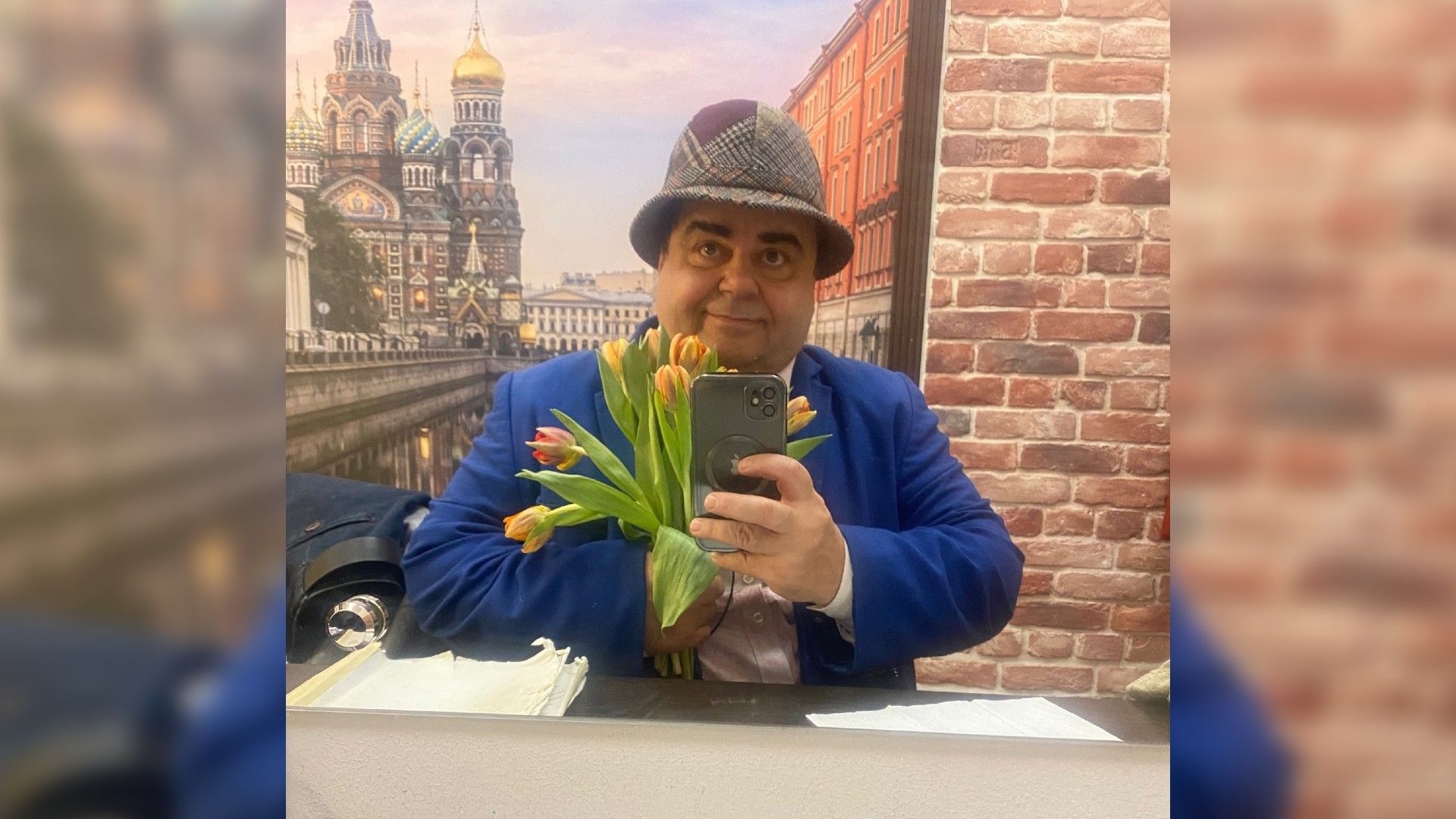 Сергей Рост в синем пиджаке, шляпе и с букетом тюльпанов в руках делает селфи на фоне храма