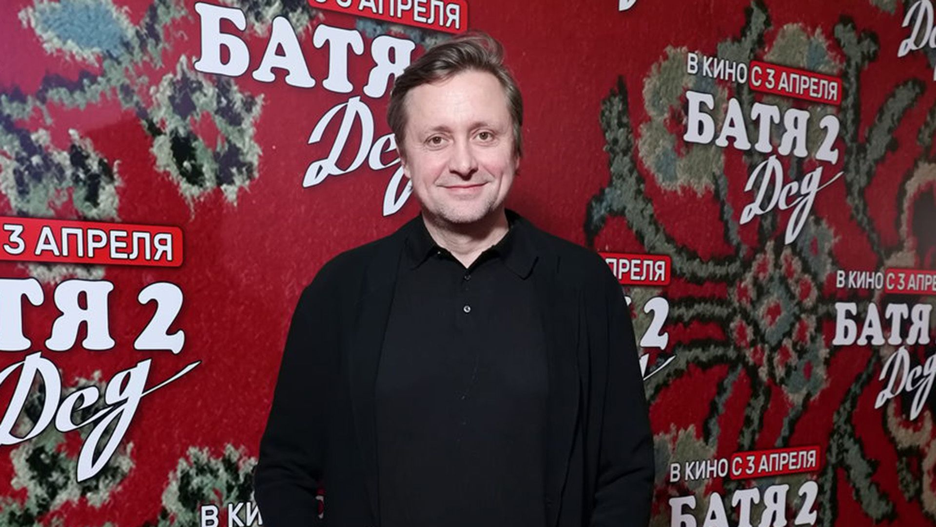 Артём Михалков в чёрной рубашке на фоне постера фильма "Батя 2. Дед"