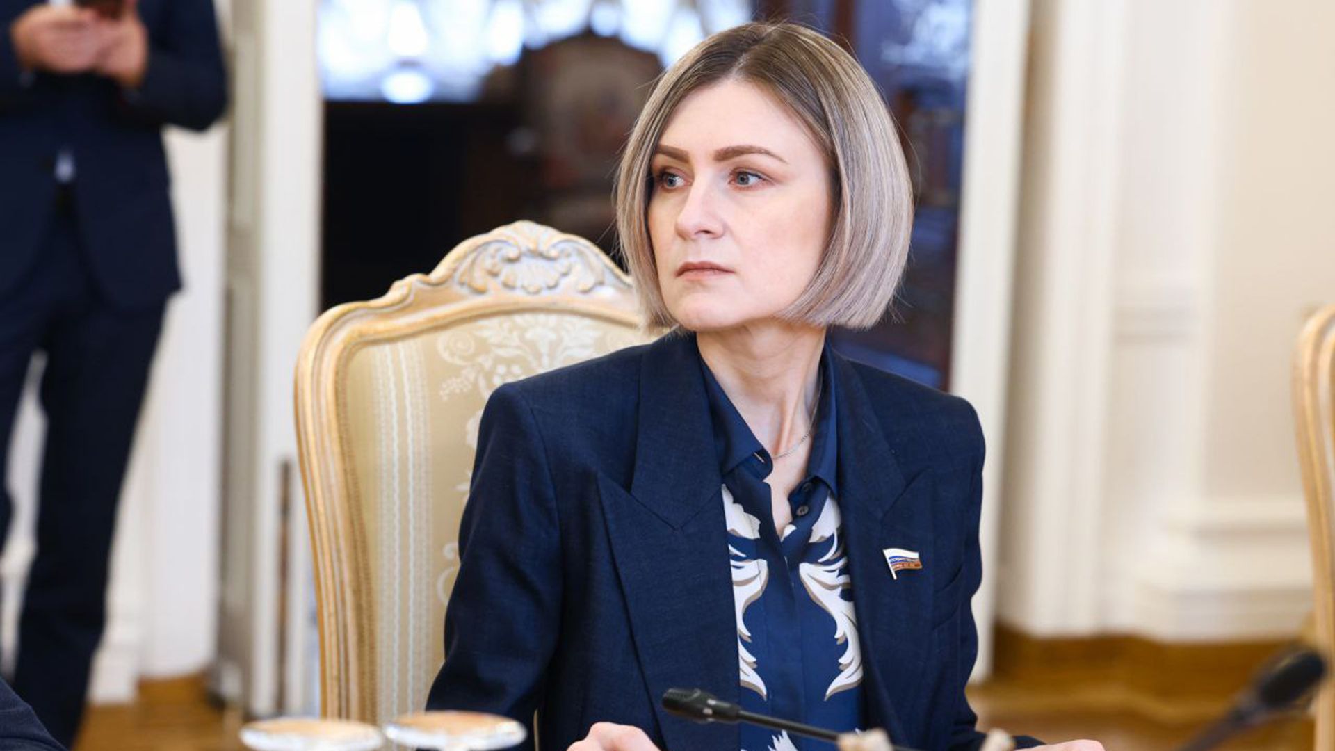 Мария Бутина в синем пиджаке сидит за столом с микрофоном