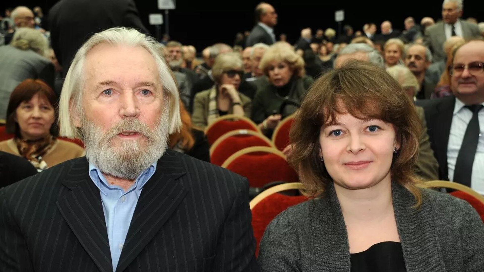 Станислав Любшин с бородой и седыми волосами и женщина среднего возраста сидят в зале среди других зрителей