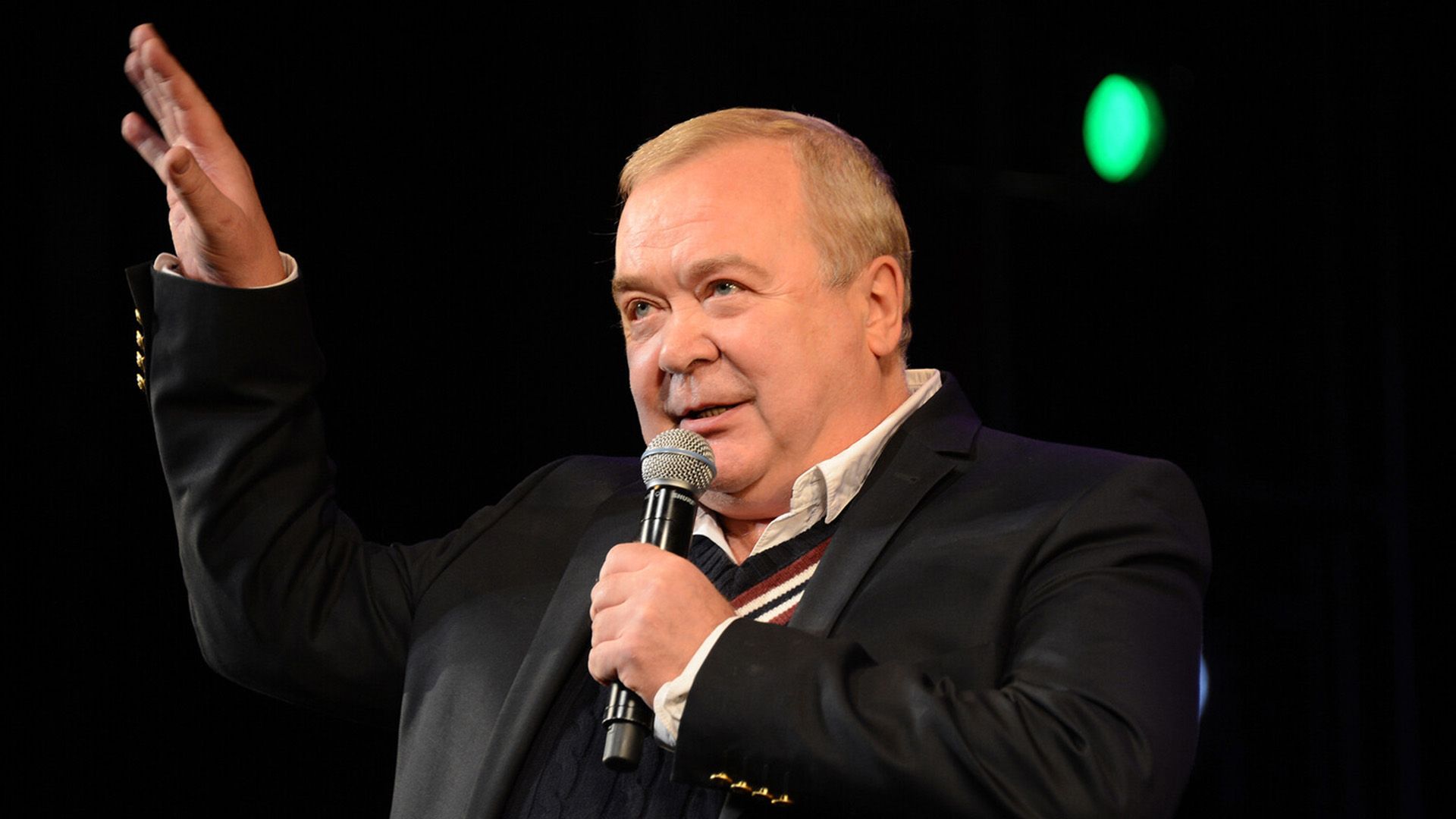 Сергей Проханов с микрофоном в левой руке и с поднятой правой