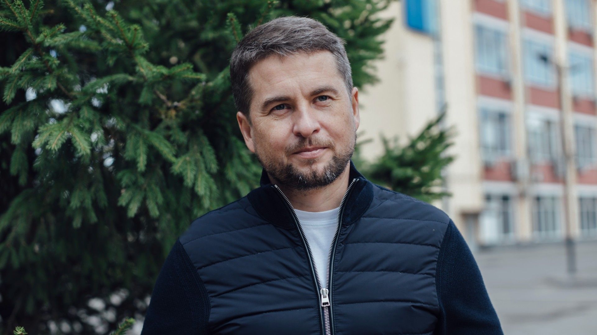 Алексей Немерюк в тёмной куртке стоит на улице у ёлки, на заднем плане здание с окнами