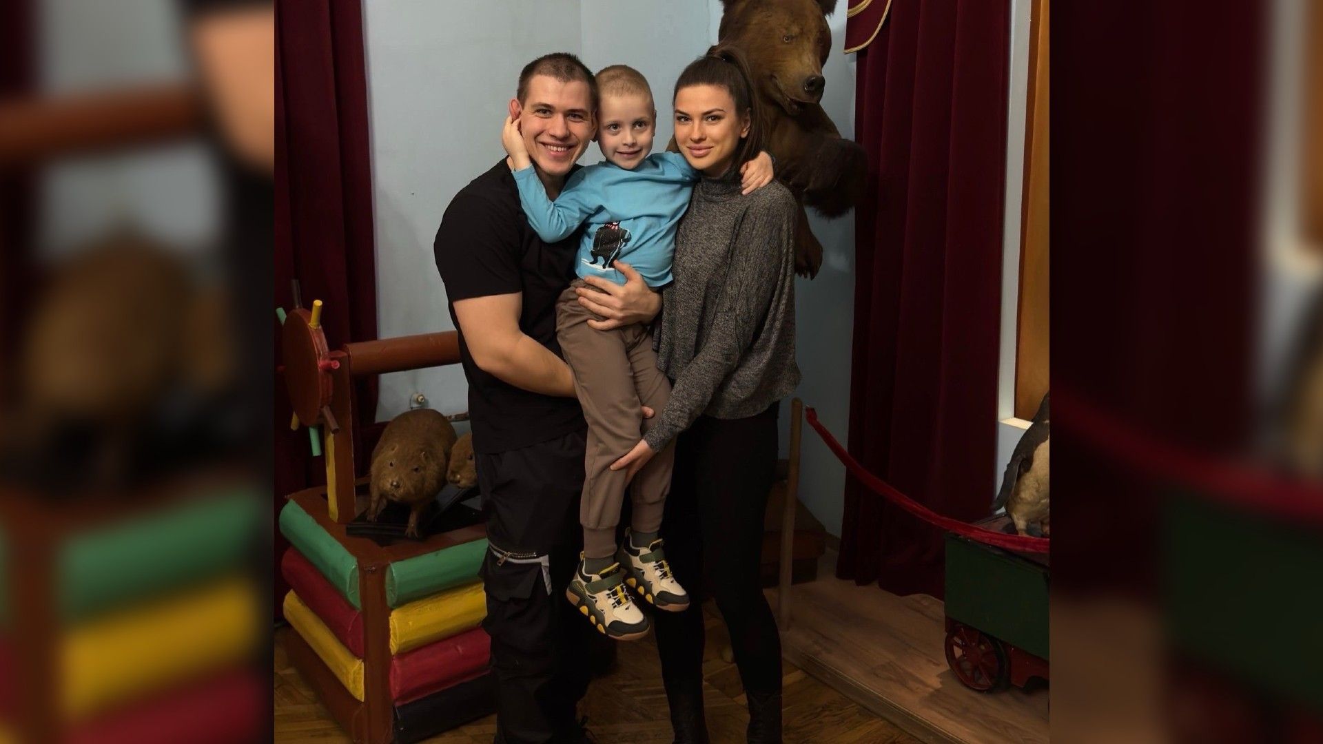 Мужчина и женщина обнимают улыбающегося мальчика на фоне чучела медведя и музейных экспонатов