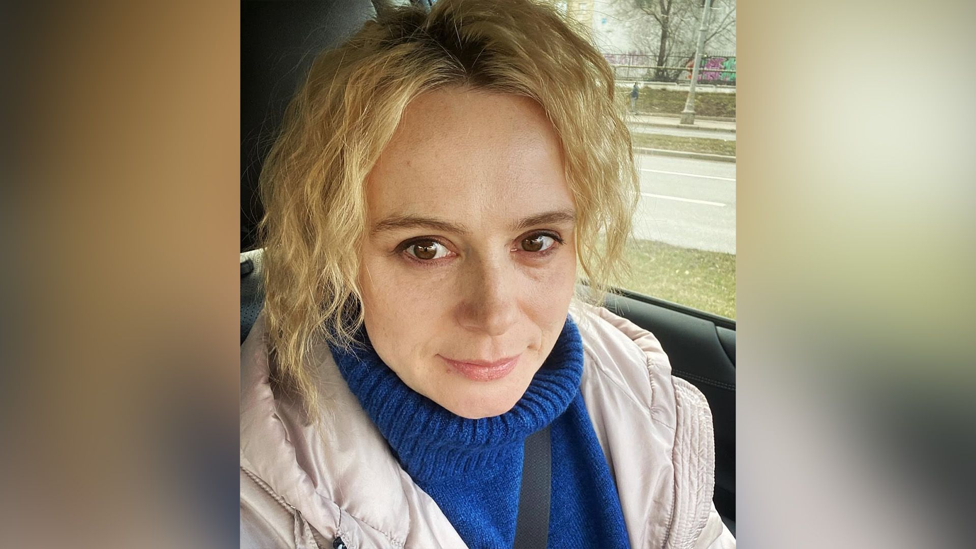 Актриса Алла Юганова в бежевой куртке и синем свитере сидит в салоне автомобиля