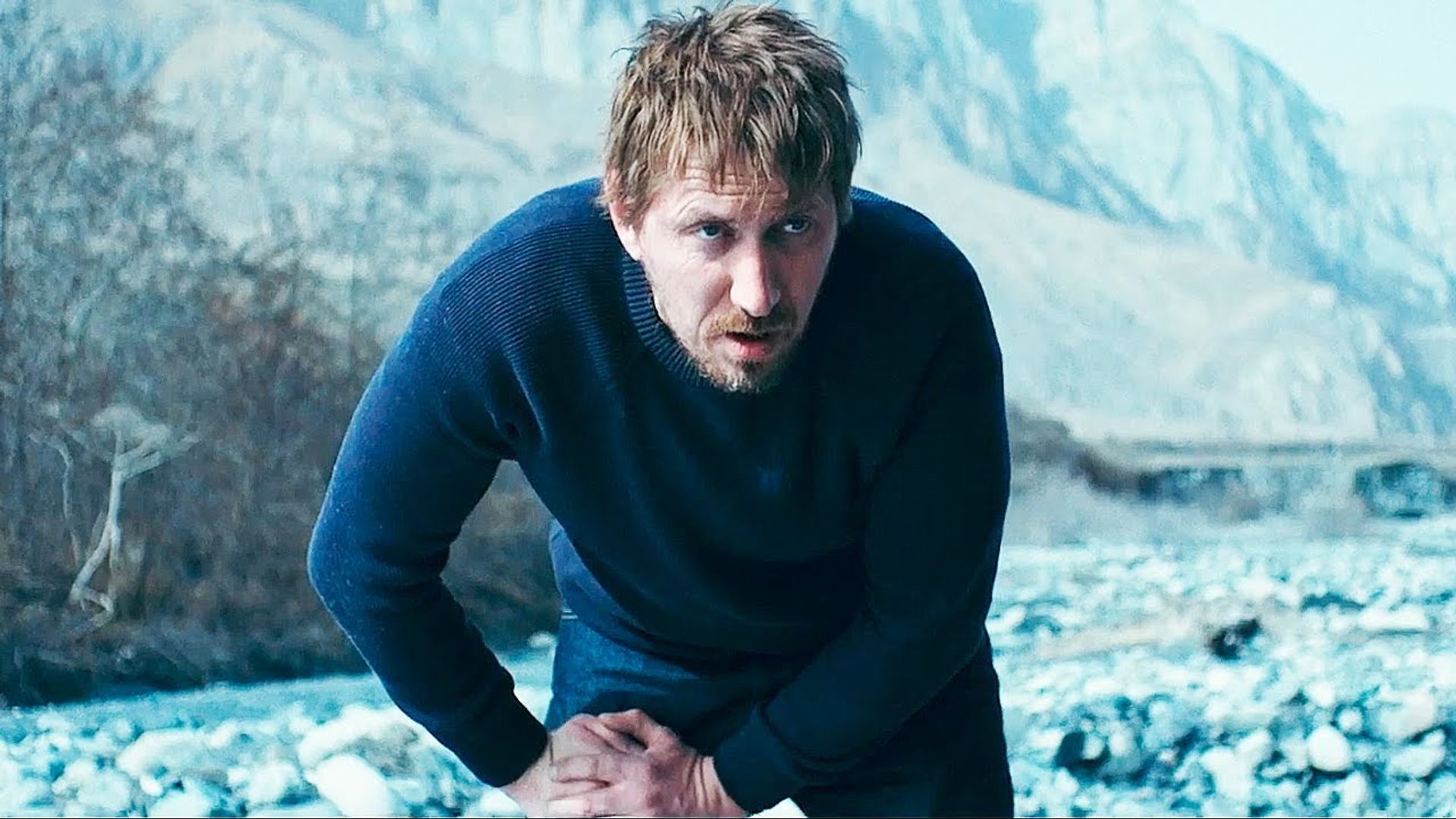 Кирилл Кяро в синем свитере склонился вперёд на фоне гор и заснеженной каменистой местности