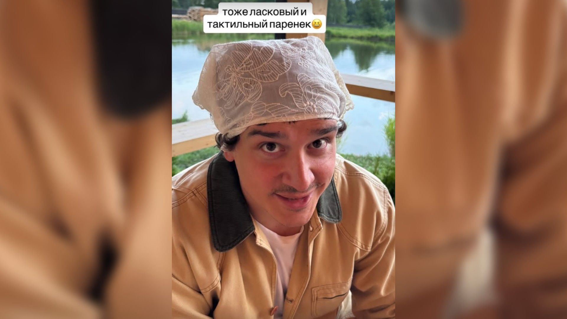 Филипп Бегак в бежевой куртке и кружевной косынке на голове смотрит вверх, за ним — водоём и зелёный пейзаж