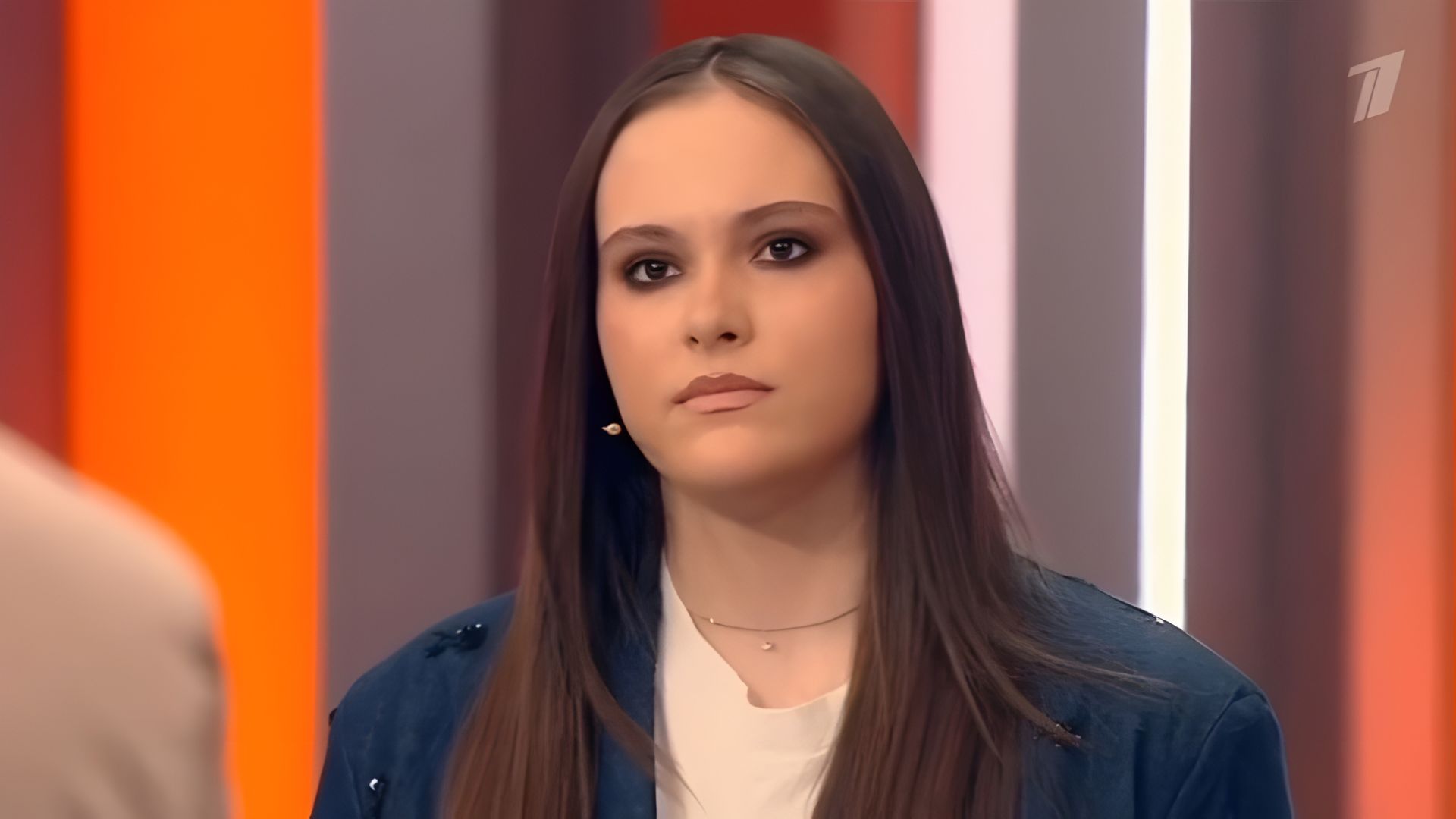 Даниэла Кравец с длинными темными волосами и серьёзным выражением лица стоит в студии ток-шоу на фоне оранжевых и белых панелей
