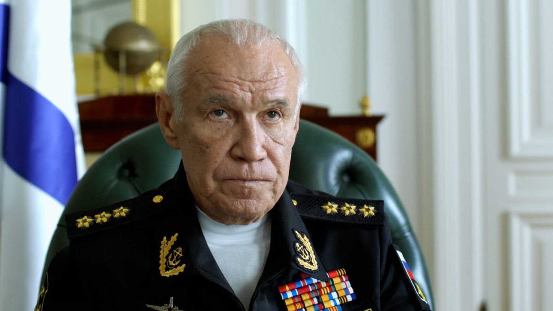 Сергей Гармаш в форме адмирала с орденами сидит на фоне флага и мраморного интерьера, глядя строго вперёд