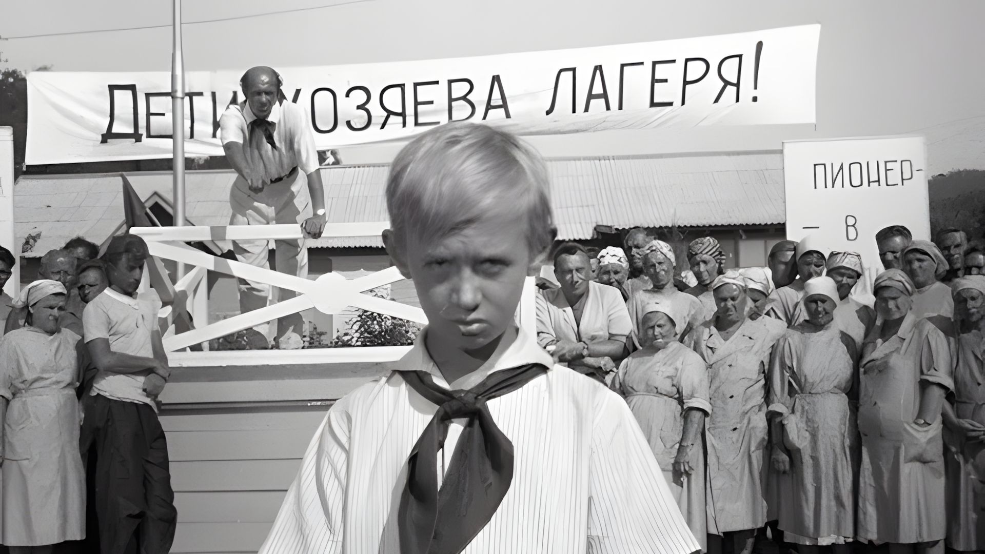 Чёрно-белое фото: мальчик в пионерском галстуке стоит на переднем плане, сзади — собрание людей и транспарант