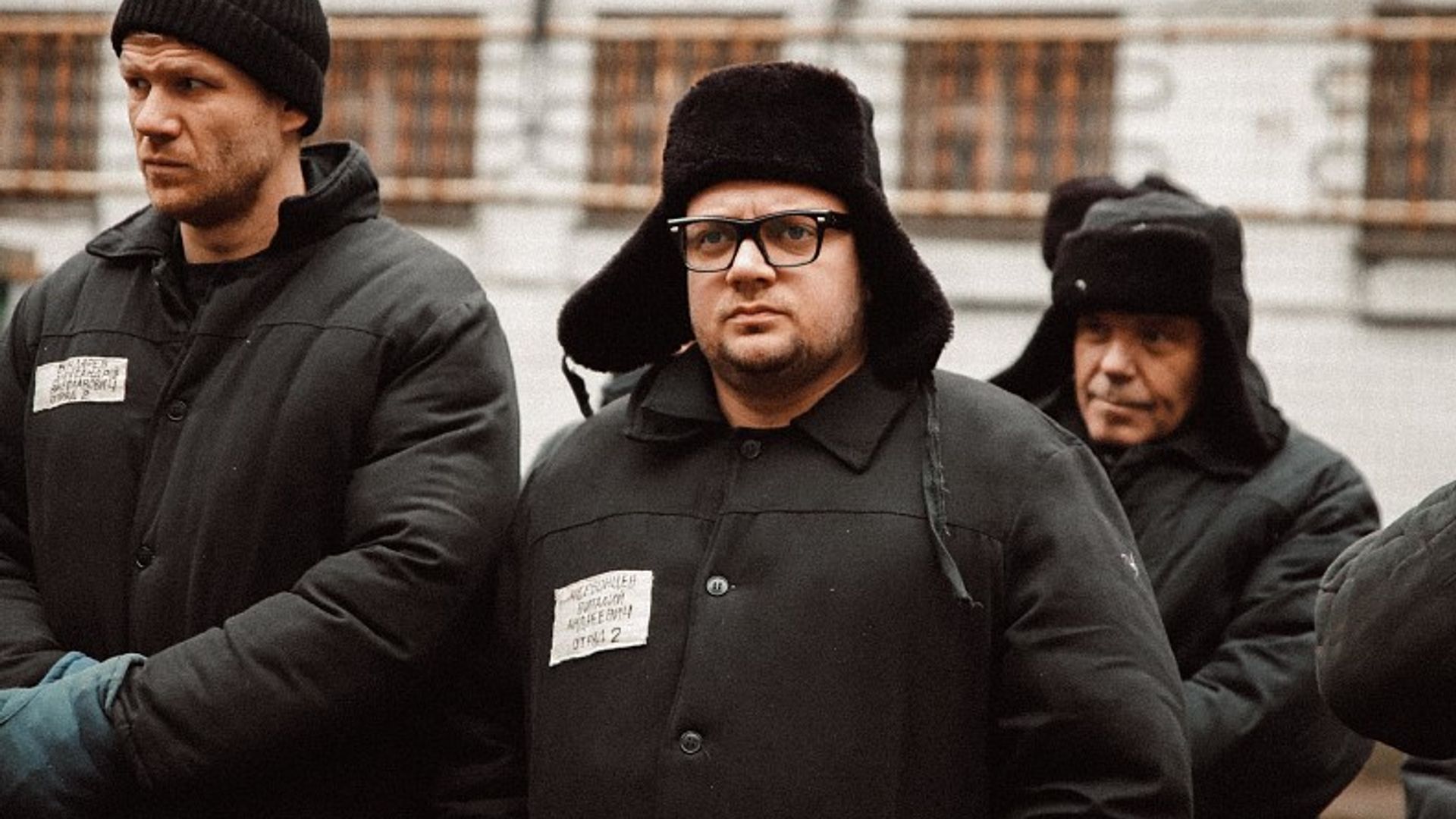 Борис Дергачев в тюремной форме и ушанках стоят в строю на фоне колонии, один из них — в очках, с серьёзным выражением лица.