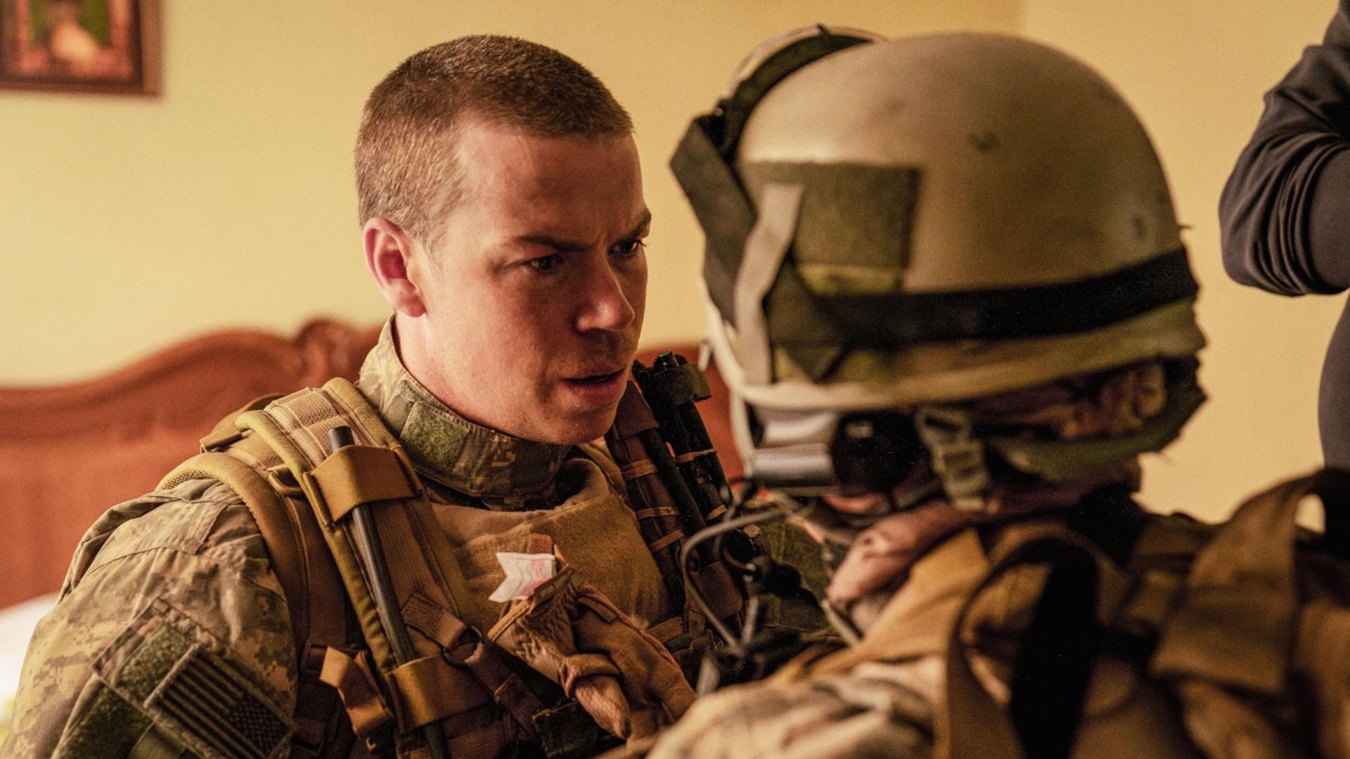 Актер Уилл Поултер в военной форме стоит около кровати и смотрит на солдата в каске