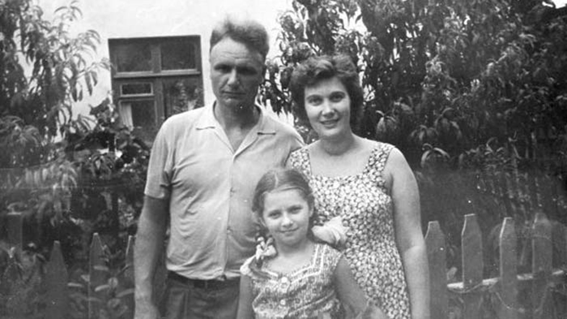 Татьяна Анциферова в детстве стоит рядом с родителями на фоне дома с садом