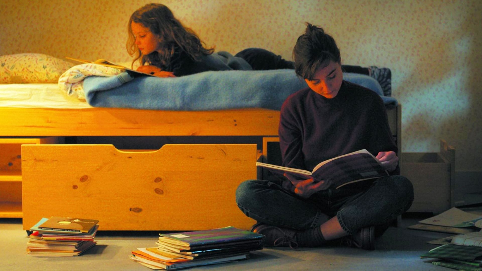 Девочка лежит на кровати и читает книгу, у кровати сидит женщина с книгой
