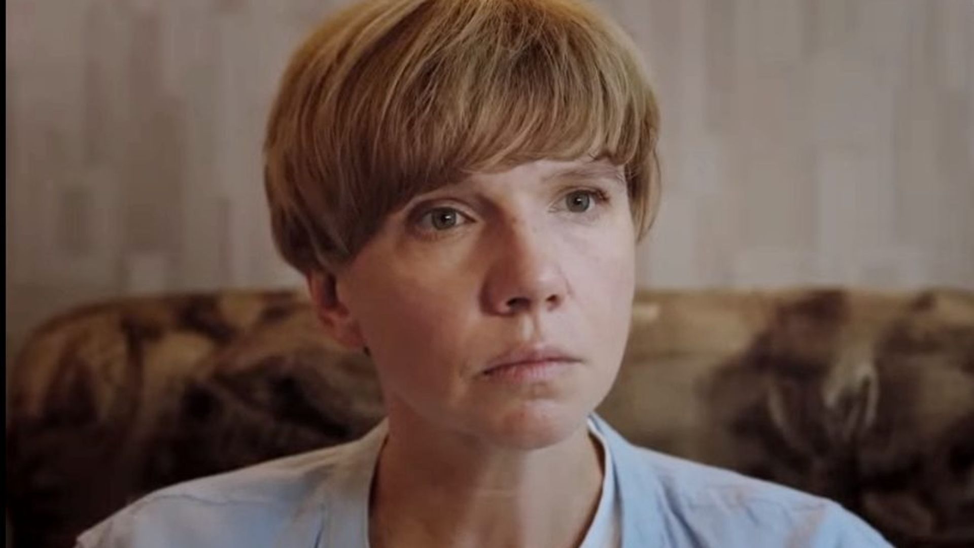 Ирина Рахманова короткой светло-русой стрижкой сидит на диване и серьёзно смотрит вперёд, сцена из фильма
