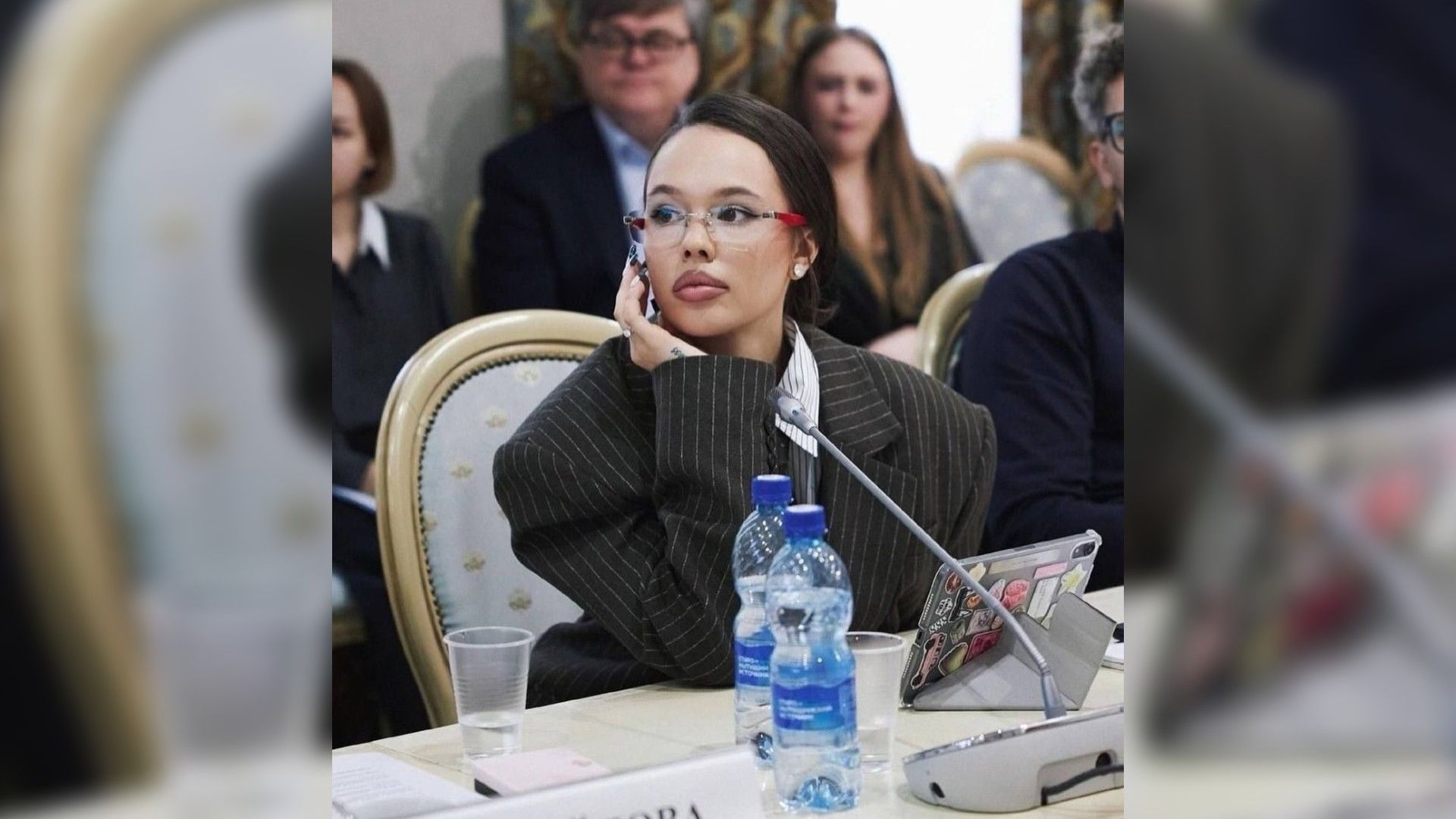 Инстасамка в очках и в полосатом пиджаке сидит за столом на совещании, опираясь на руку, перед ней планшет и бутылки воды