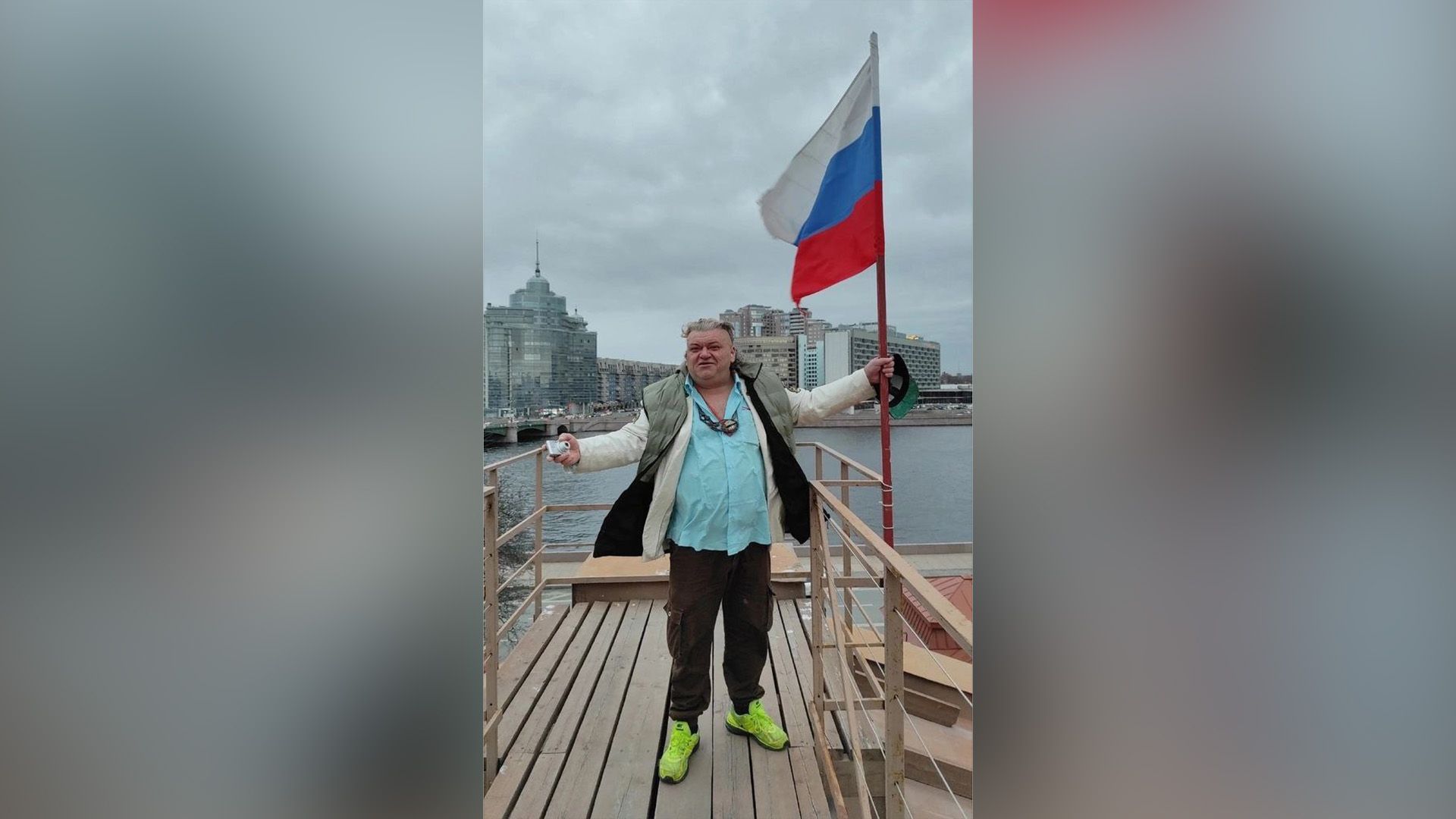 Пророк Санбой стоит на набережной, держа в руке большой флаг РФ