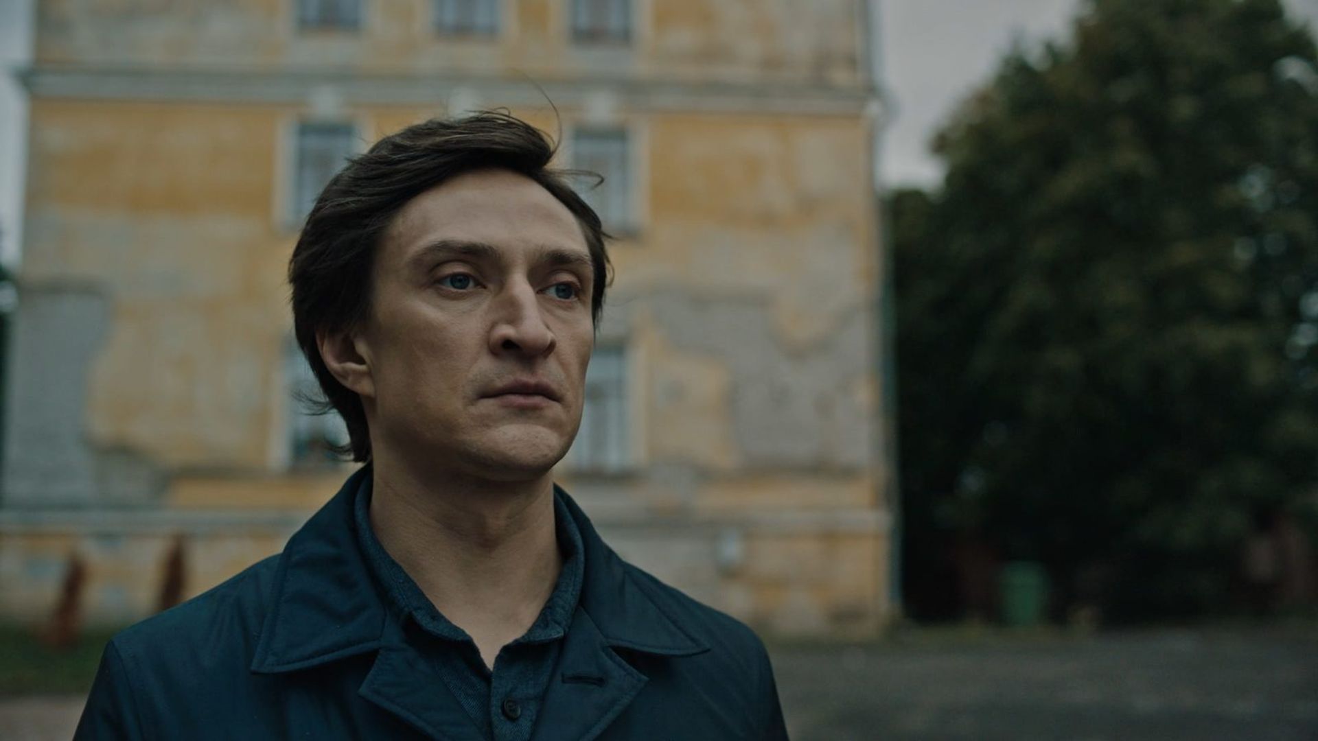 Юрий Чурсин c серьёзным выражением лица стоит на улице на фоне старого облупившегося здания