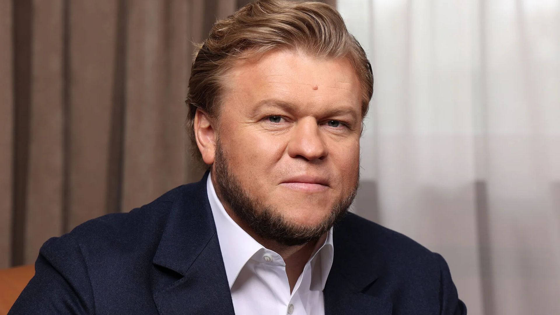 Морозов Денис в темно-синем пиджаке и белой рубашке сидит в помещении, глядя в камеру с лёгкой улыбкой