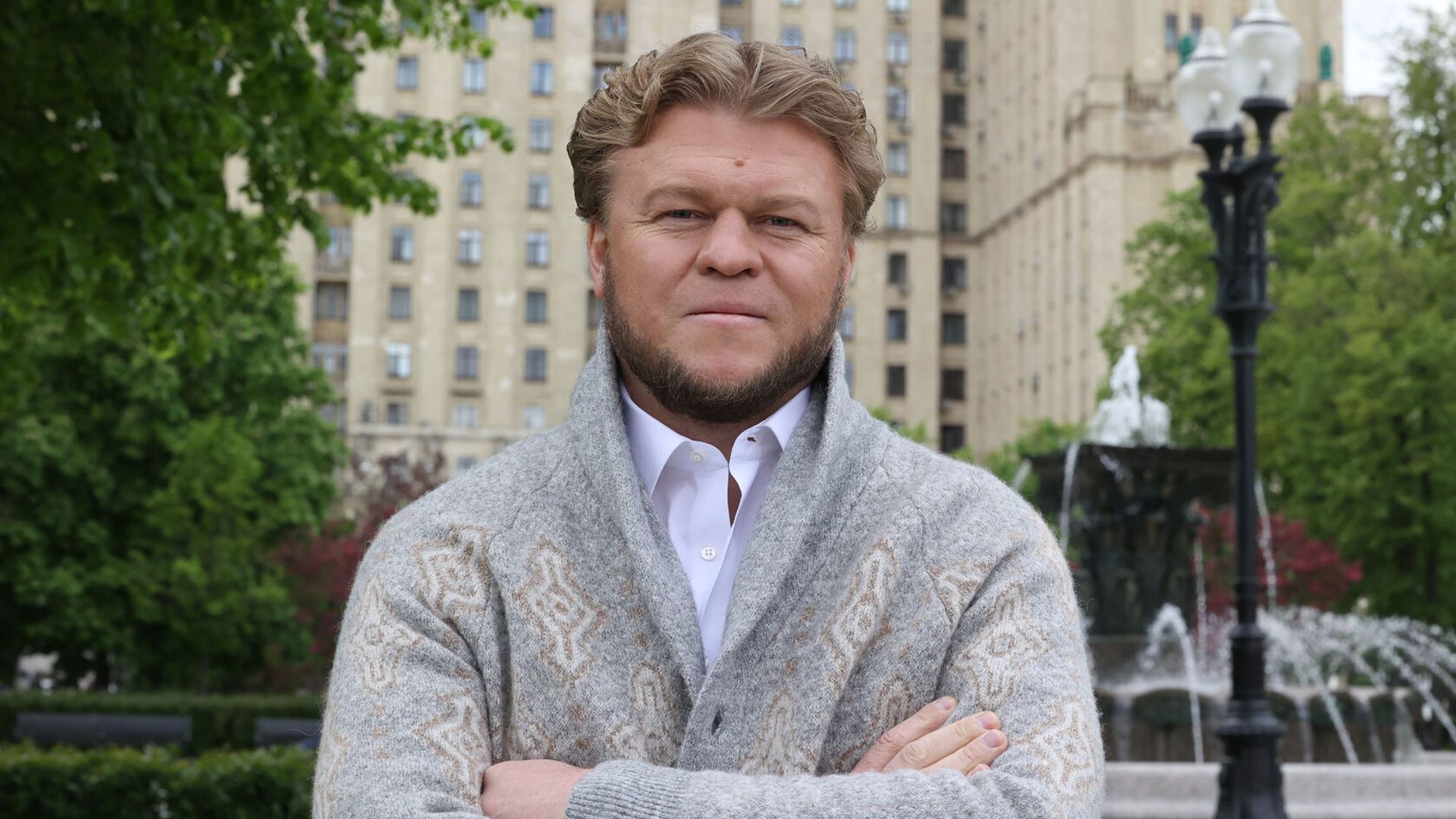 Денис Морозов в сером вязаном кардигане стоит на улице с фонтаном и многоэтажным зданием на заднем плане