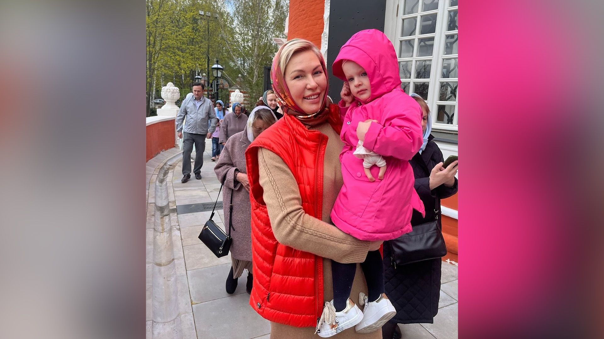 Наталья Рагозина в платке и красном жилете держит на руках дочь, одетую в розовую куртку