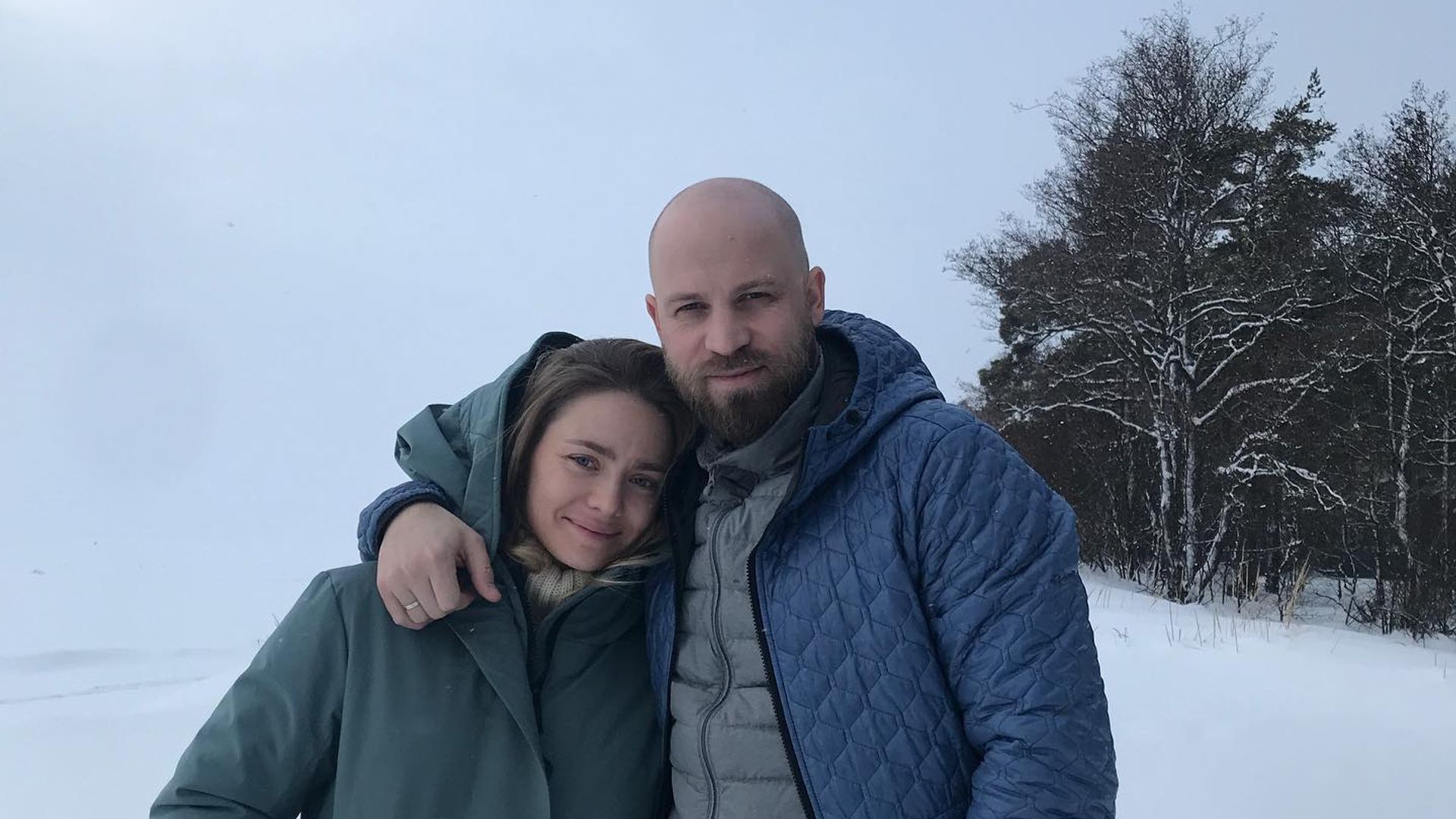 Карина Разумовская и Егор Бурдин в зимних куртках стоят обнявшись на фоне заснеженного пейзажа и деревьев