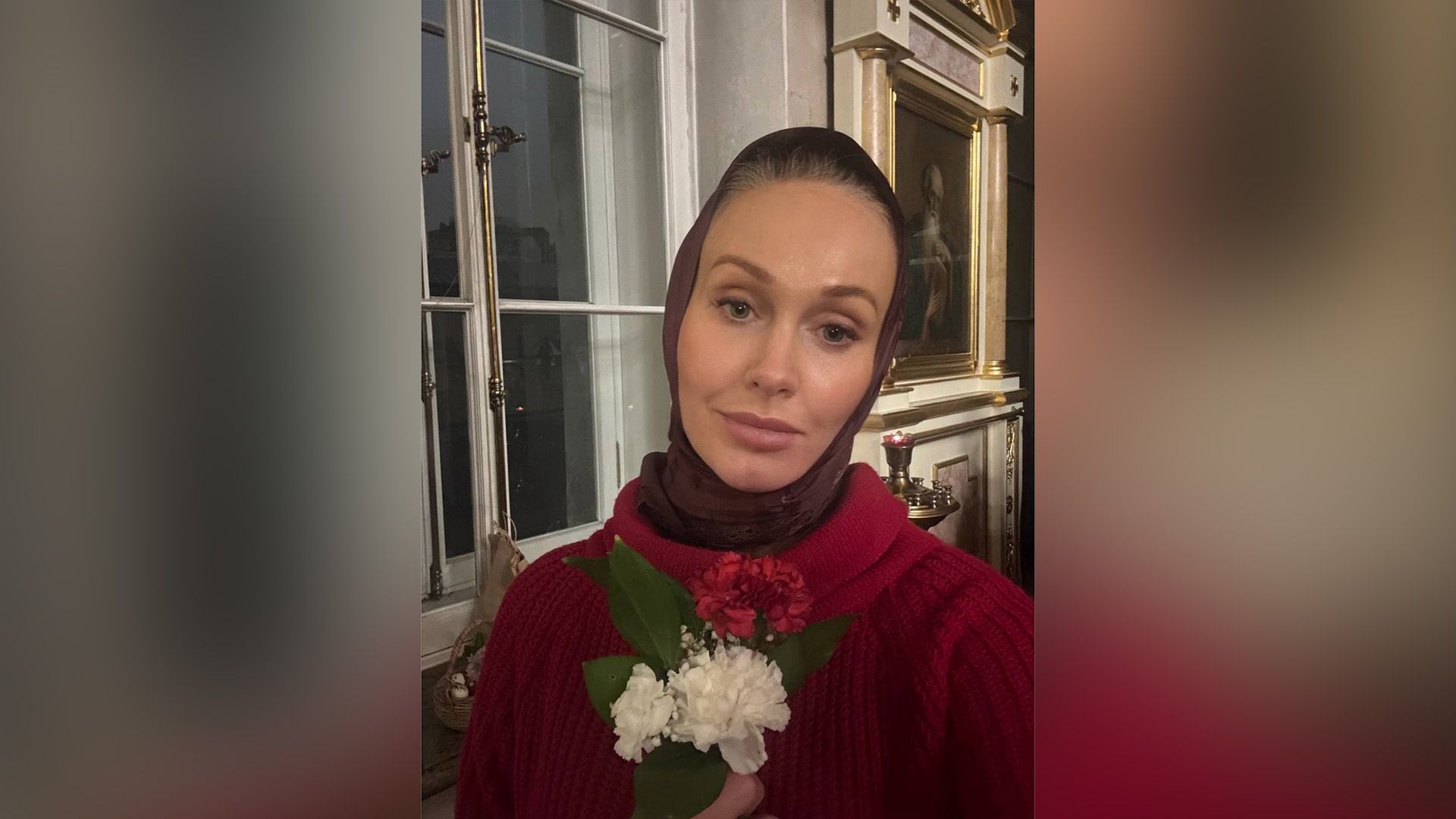 Полина Невзорова в красном свитере и платке держит в руках белые цветы в интерьере церкви