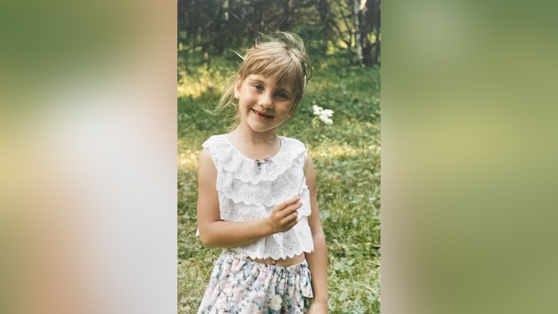 Дарья Пицик в детстве, одетая в белую майку и юбку, стоит на зелёной лужайке, держа в руке белый цветок