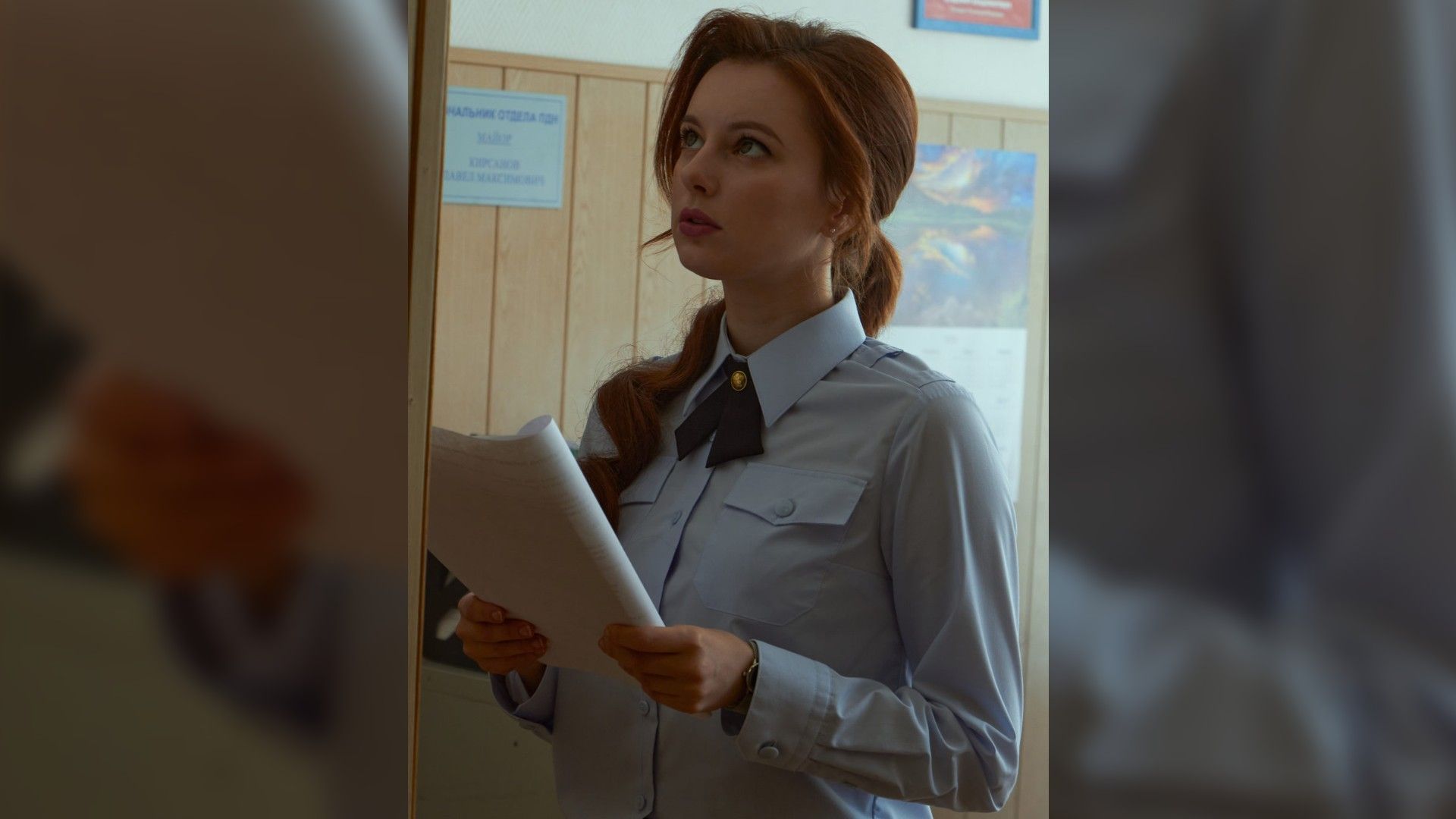Маруся Климова в голубой форме с бумагами в руках стоит в помещении и смотрит вверх