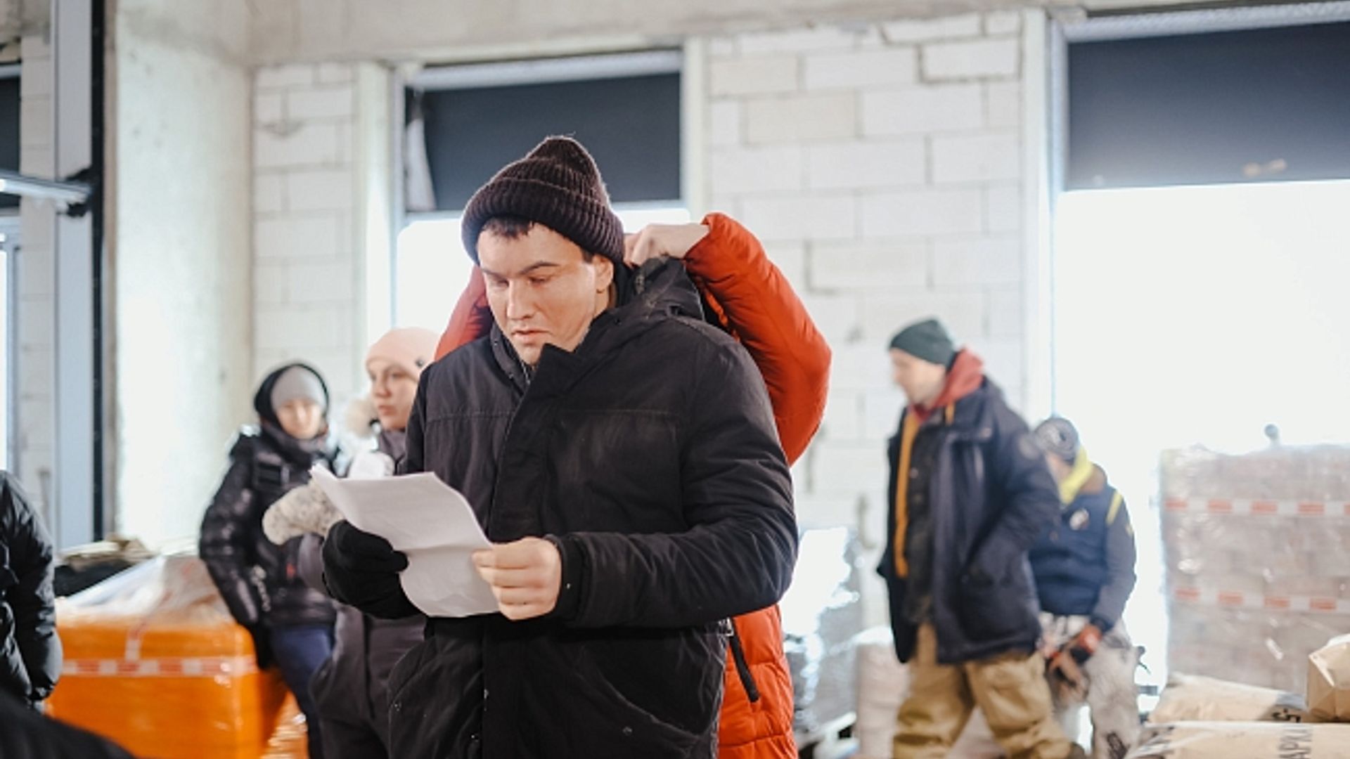 Павел Прилучный в черной куртке и вязаной шапке читает лист бумаги в помещении, вокруг люди в зимней одежде