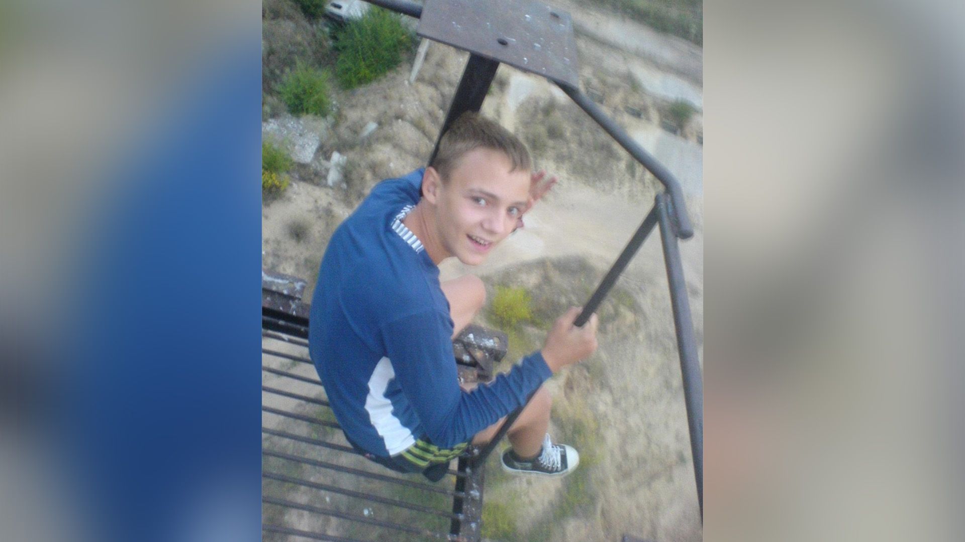 Максим Тригубчак сидит на железной конструкции, свесив ноги, и машет рукой в камеру