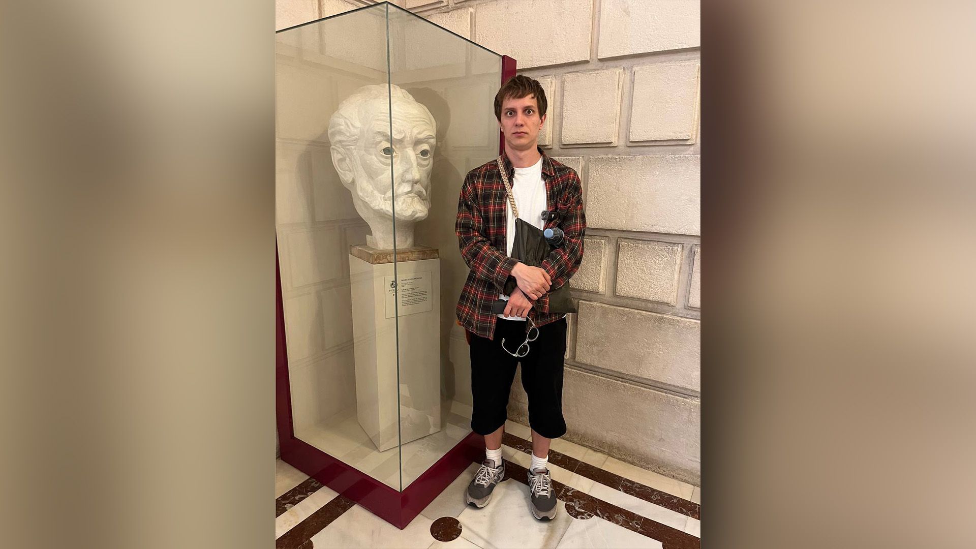 Андрей Айрапетов в клетчатой рубашке стоит возле мраморного бюста в музее
