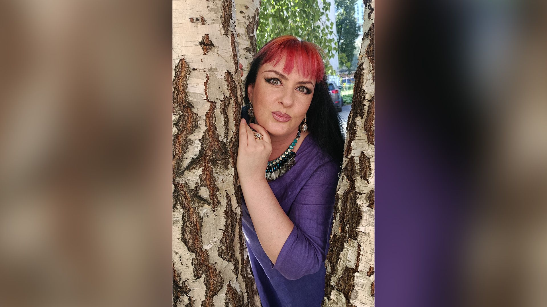 Наталья Шоколадкина в фиолетовом платье выглядывает между двух берёз